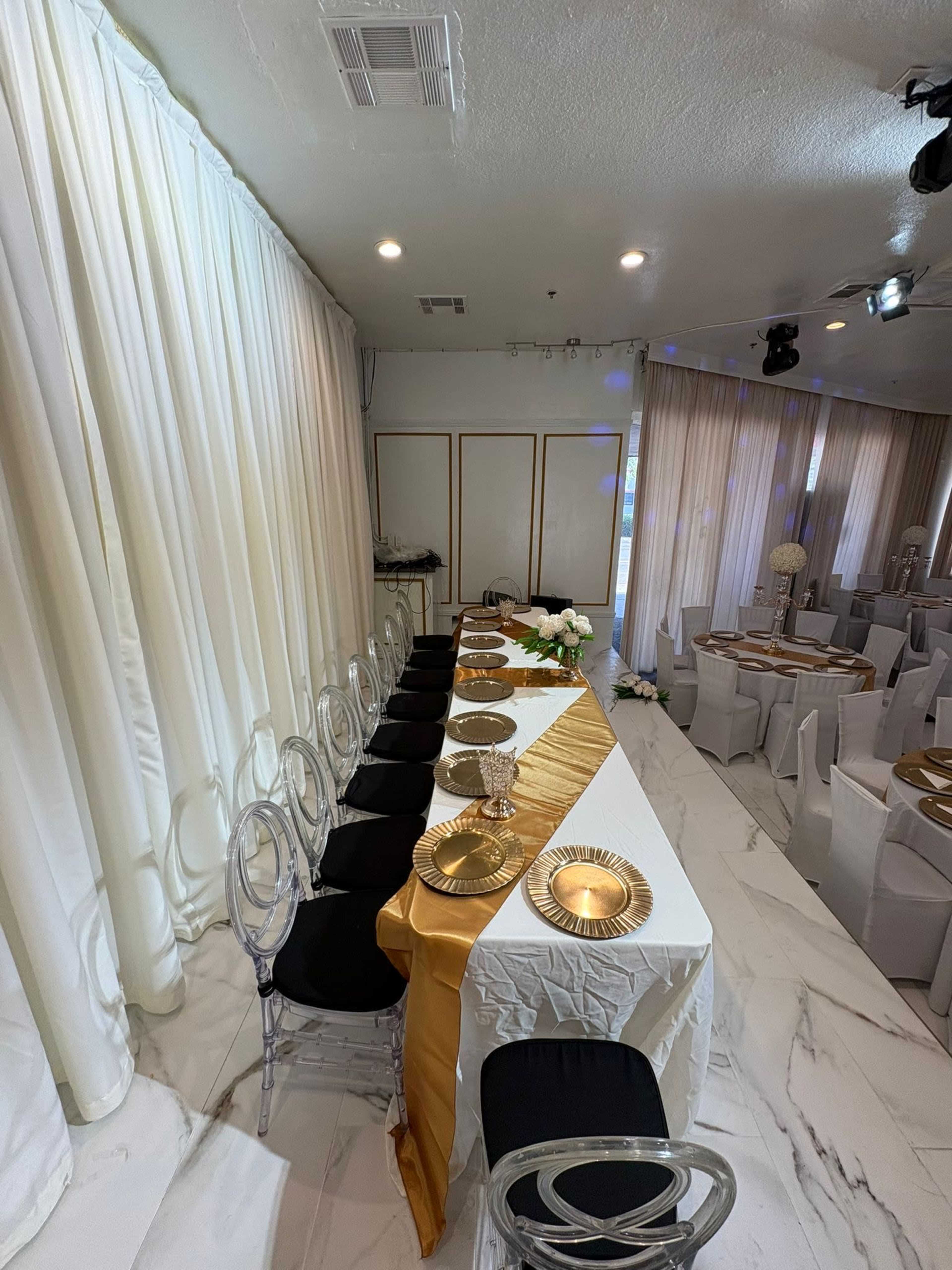 A long table is set with gold plates and a gold runner, surrounded by clear acrylic chairs in a room with white drapery and soft lighting.
