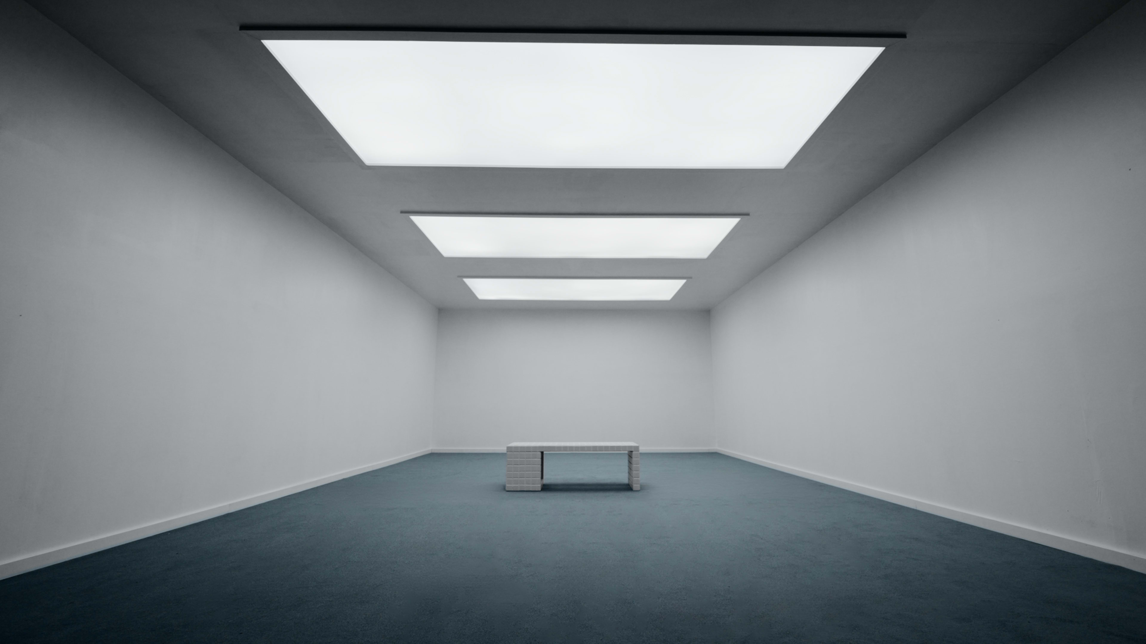 A sparsely furnished room features a white table centered under three skylights illuminating the space.
