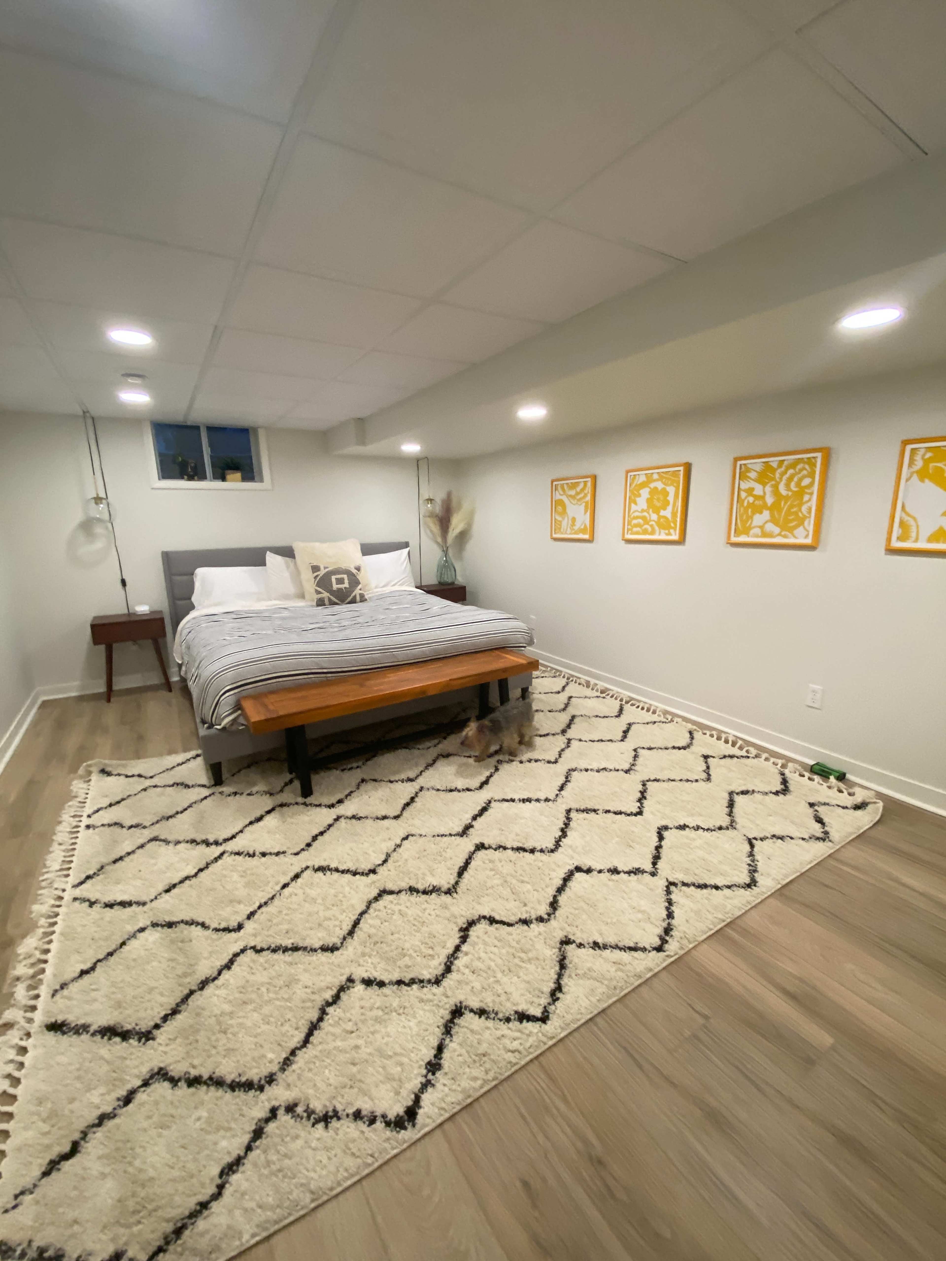 The image shows a bedroom with a bed on a rug, complemented by wooden furniture and framed yellow art on the walls.