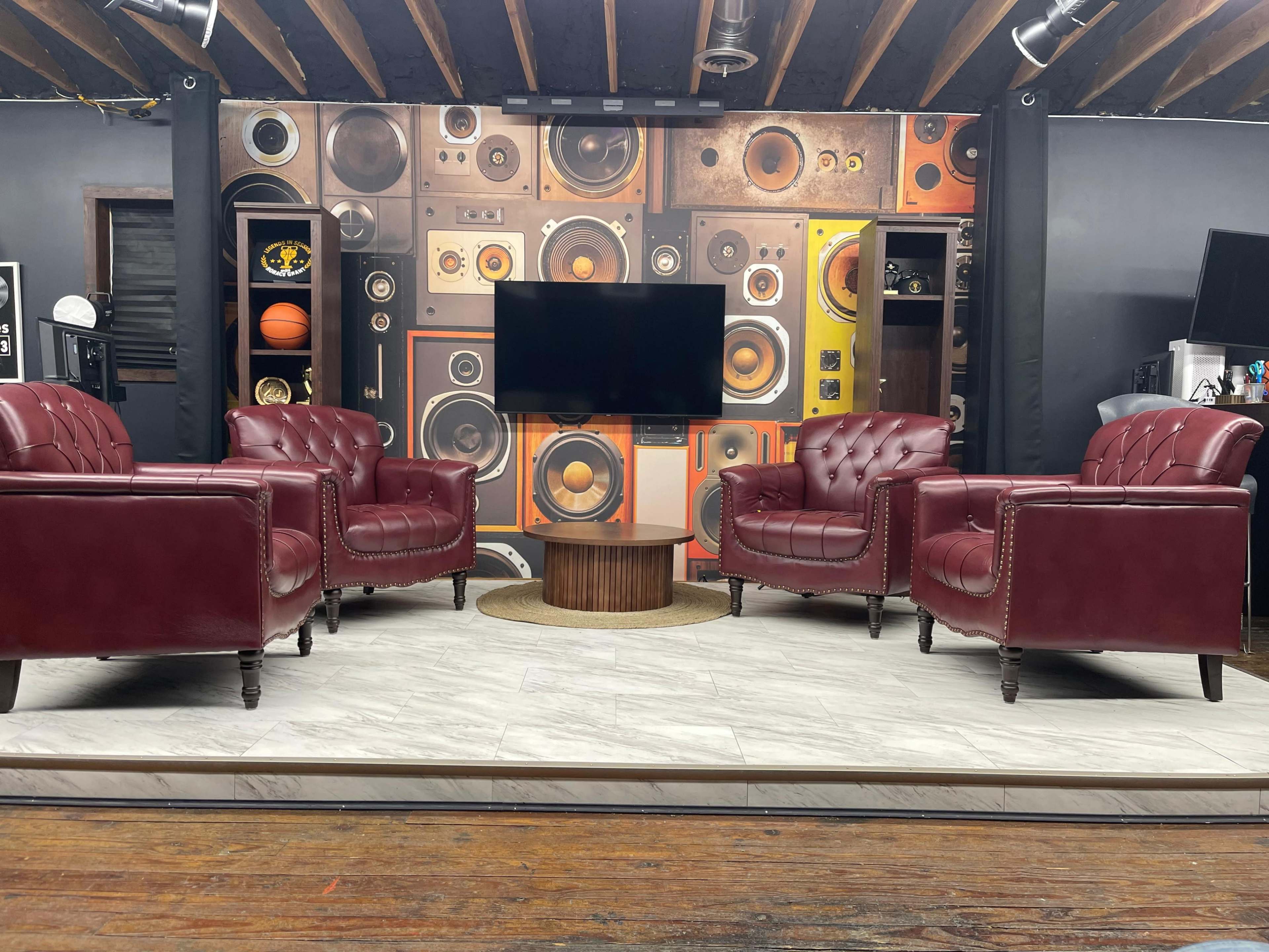 The image shows a room with four burgundy leather chairs arranged around a circular wooden table, set against a wall decorated with colorful speaker graphics and a television.