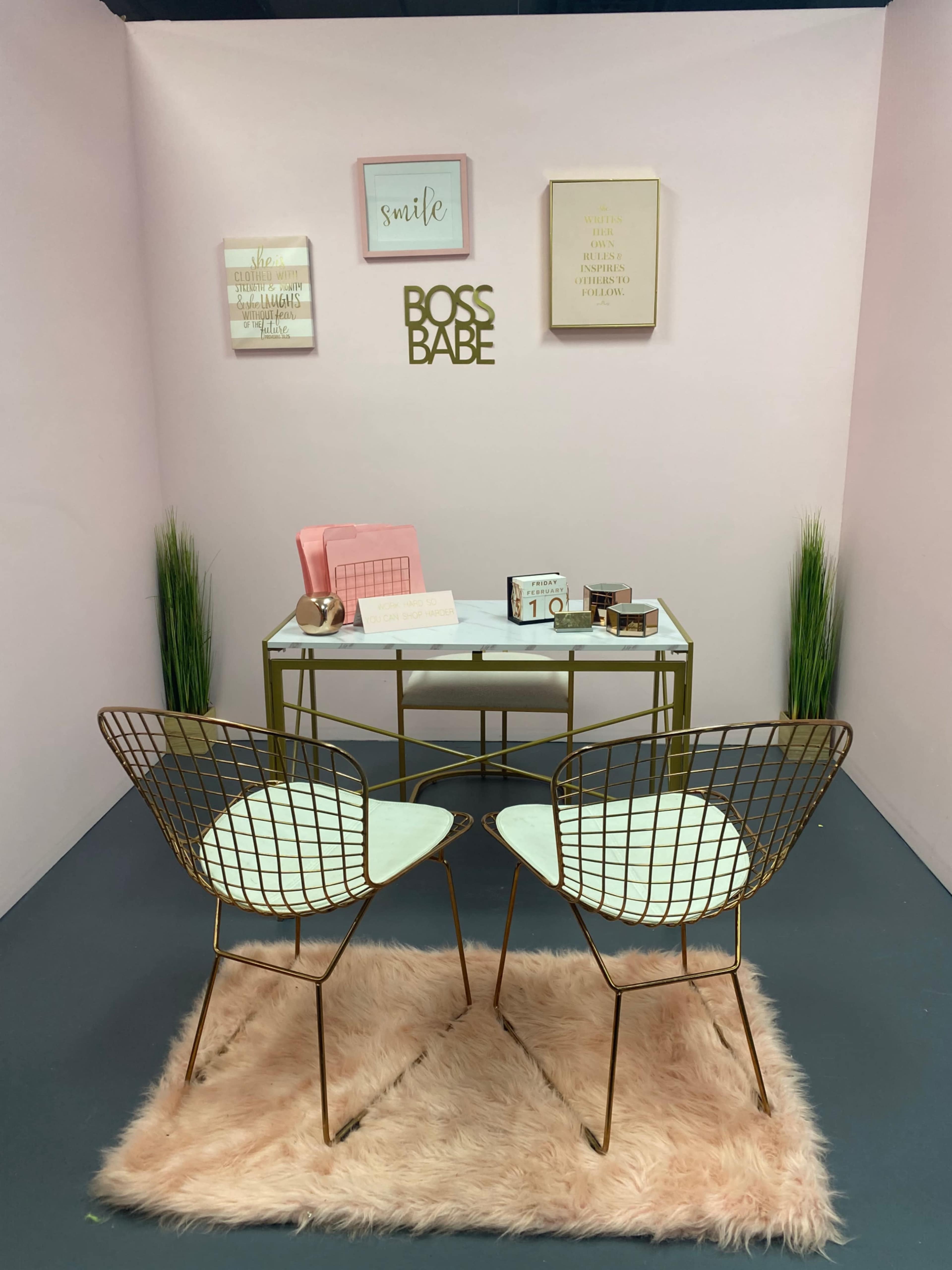 The scene features a small office space with a gold and white desk, two modern wire chairs, and decorated pink walls displaying motivational posters.