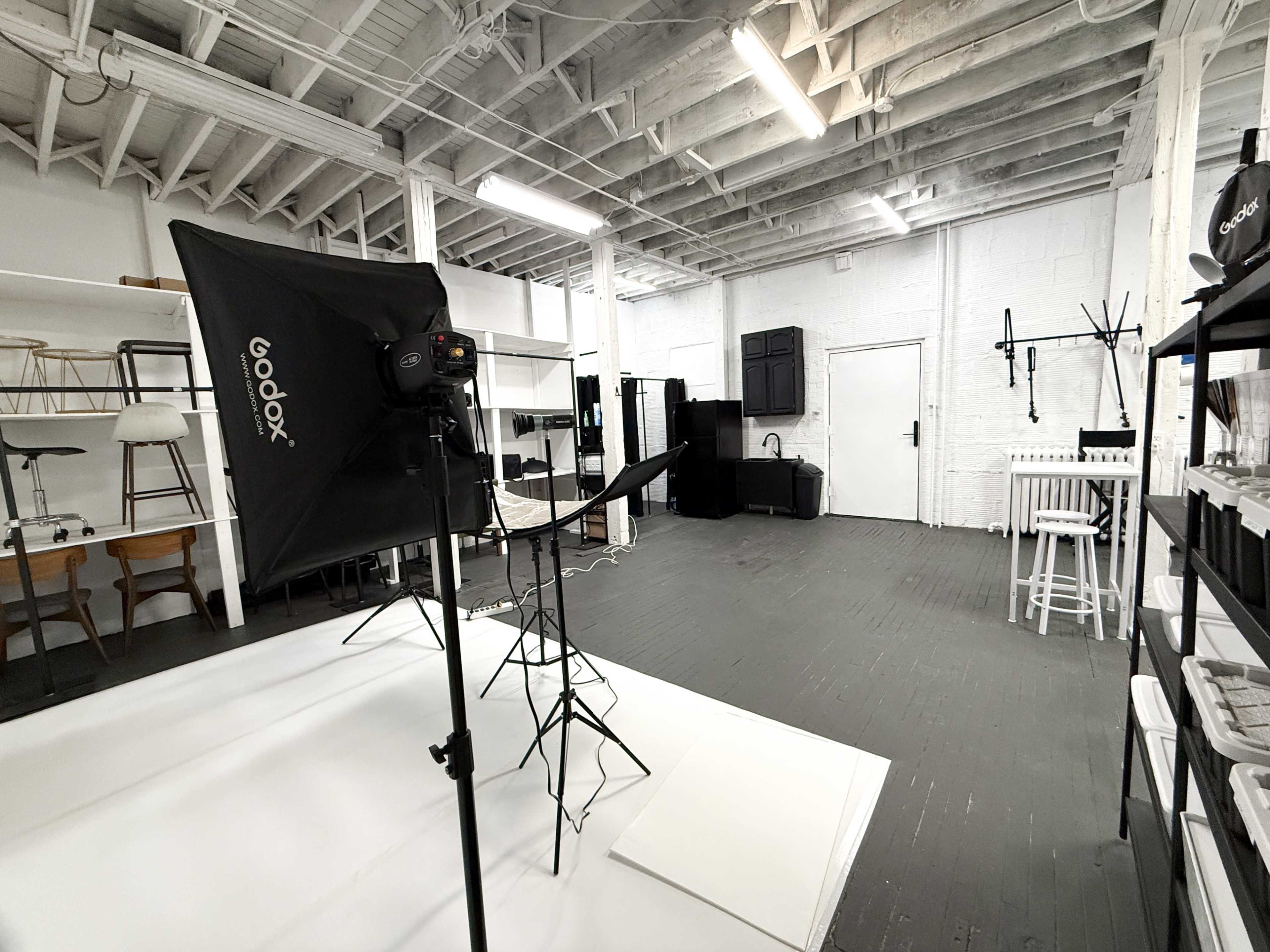 The image shows a photography studio with lighting equipment set up, including a large softbox, surrounded by various furniture and backdrops.
