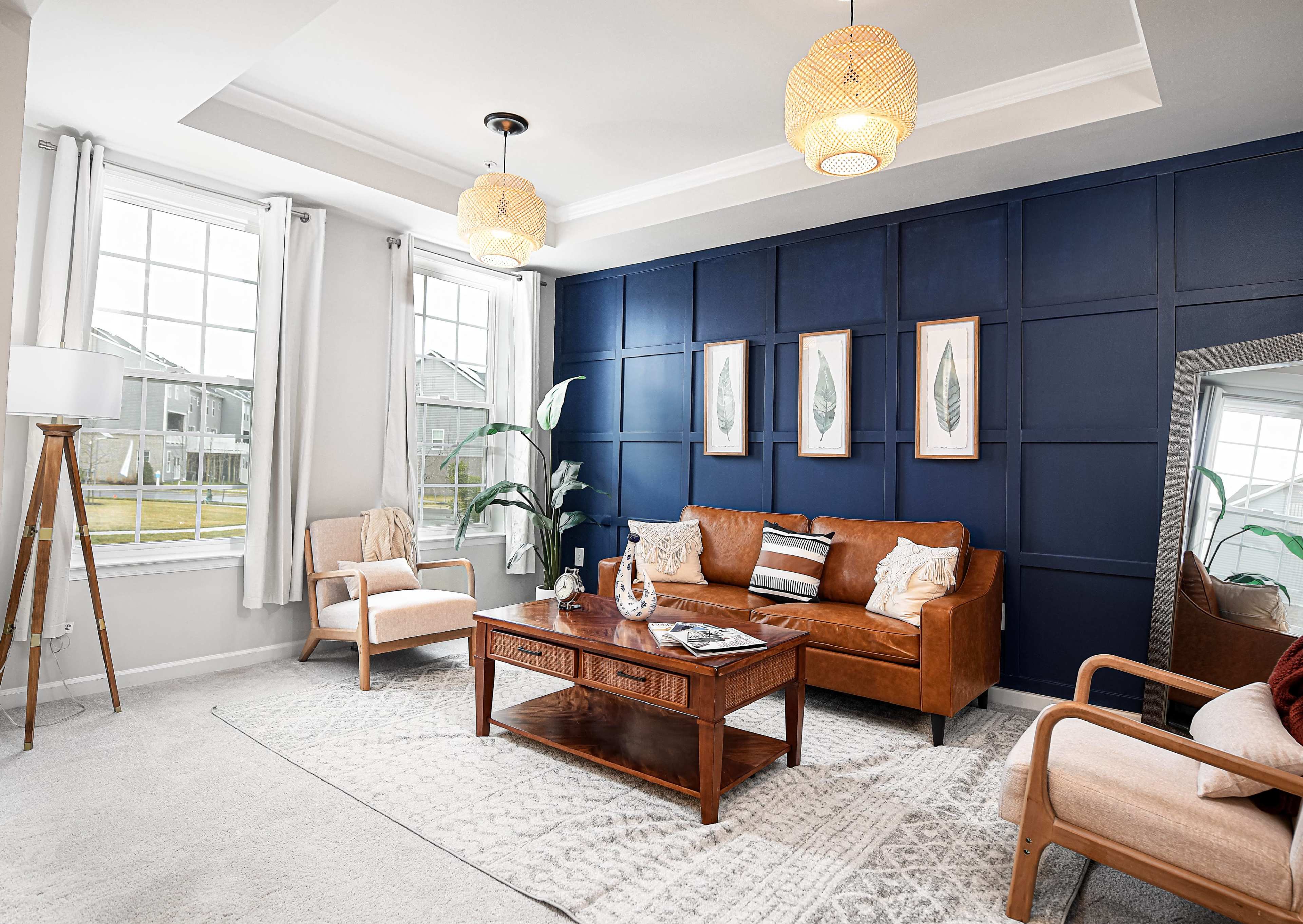 The living room features a brown leather sofa, two armchairs, a wooden coffee table, and wall art, set against a navy blue accent wall, with large windows letting in natural light.