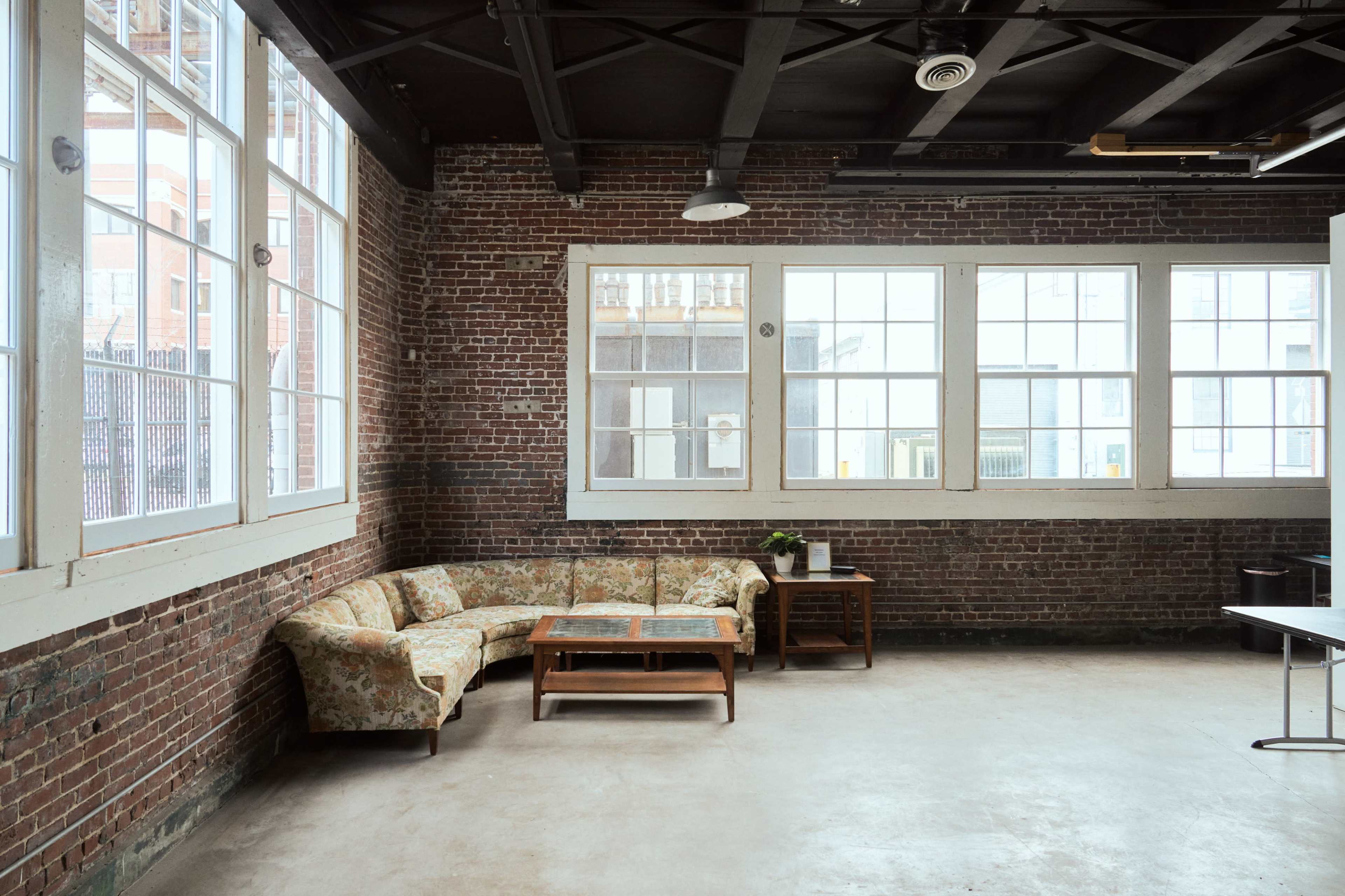 A spacious room with exposed brick walls, large windows, a floral-patterned couch, and a wooden coffee table.