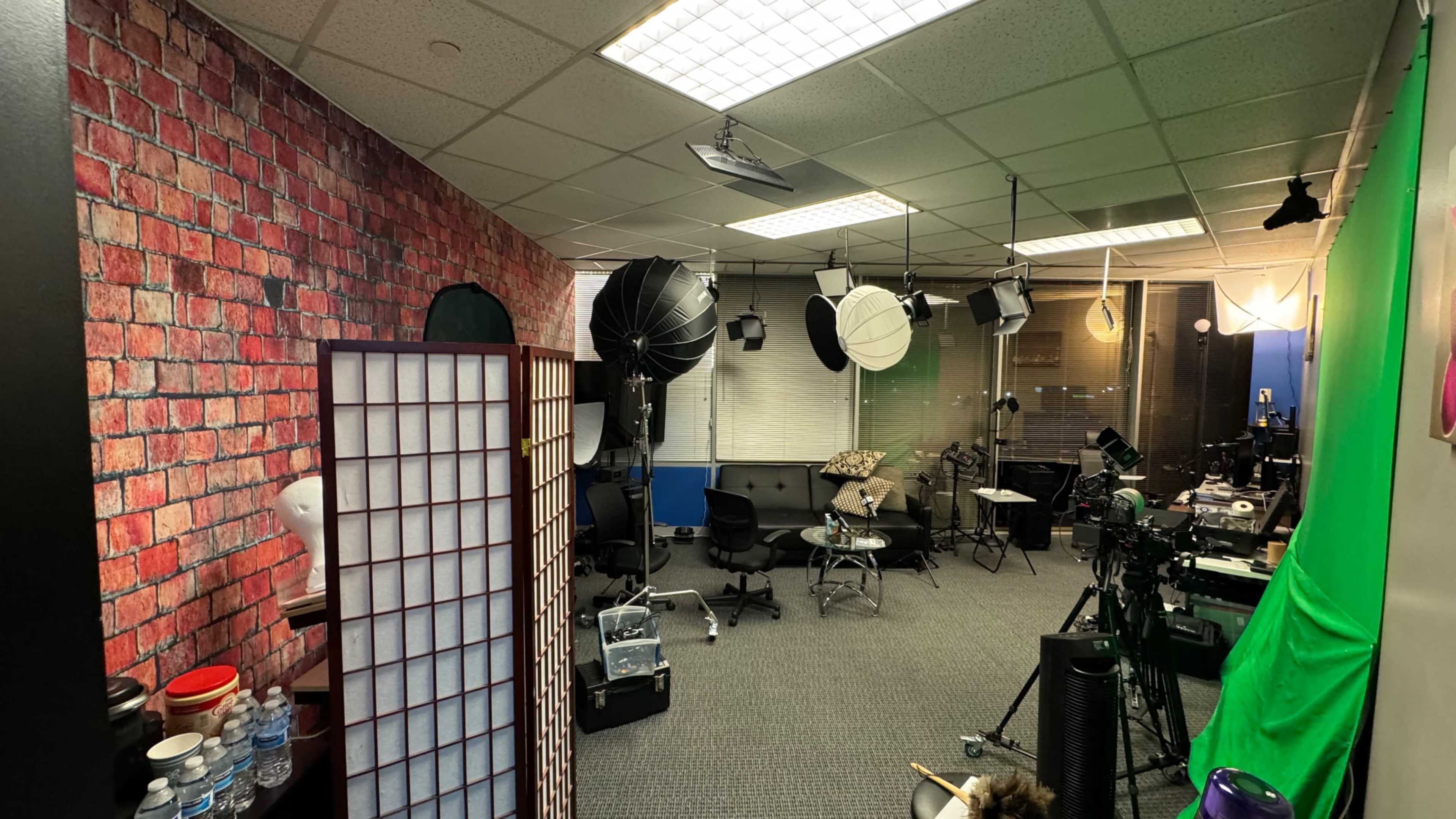 A studio setup with a brick-patterned wall, lighting equipment, chairs, and a camera on a tripod in a well-lit space.