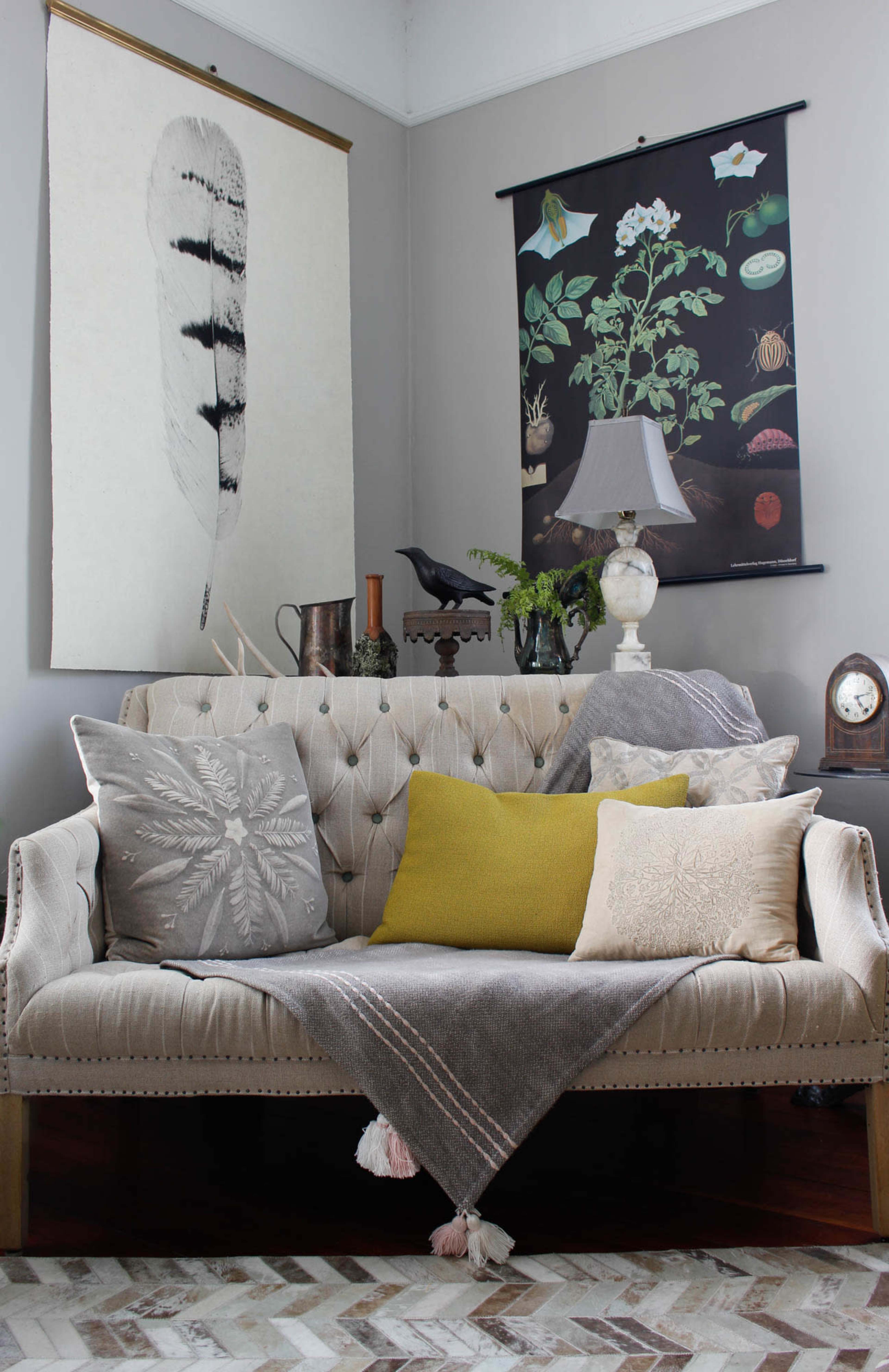 A tufted beige sofa adorned with various cushions and a throw is positioned against walls featuring botanical posters and a table lamp.
