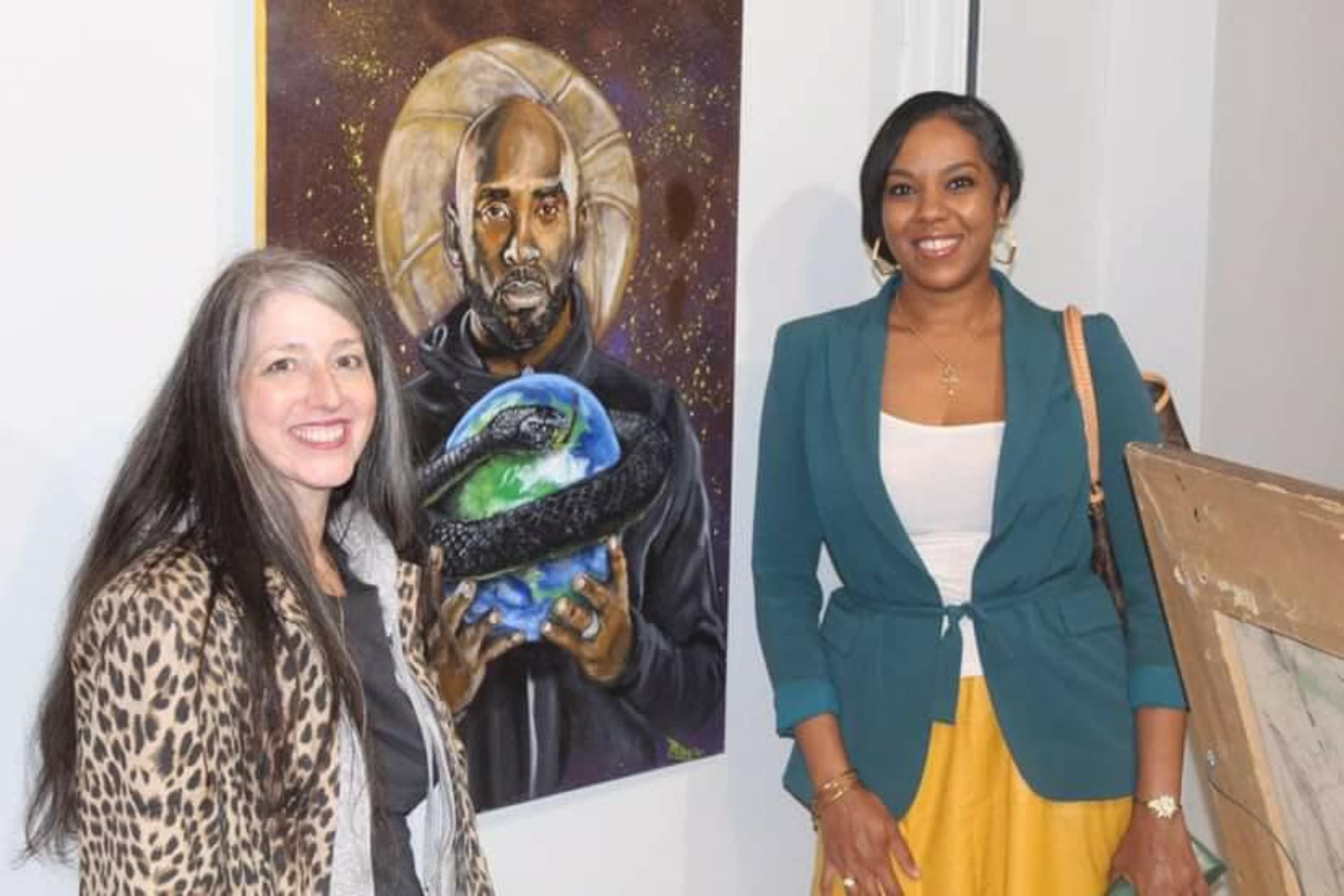 Two women stand in front of a colorful painting depicting a man holding a globe, with one woman wearing a patterned jacket and the other in a blazer and skirt.