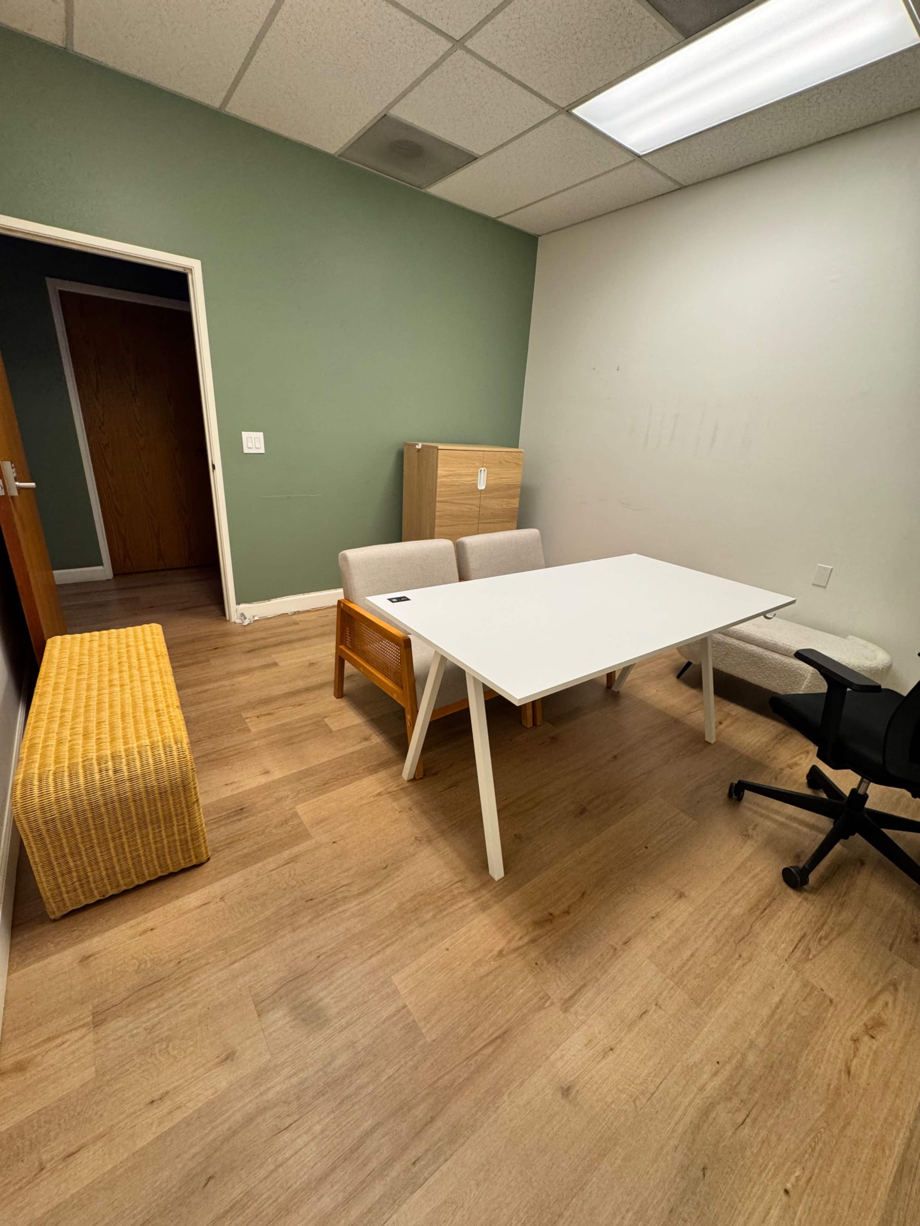 The image shows a small office space featuring a white table, two chairs, a bench, and a storage cabinet against a green wall.