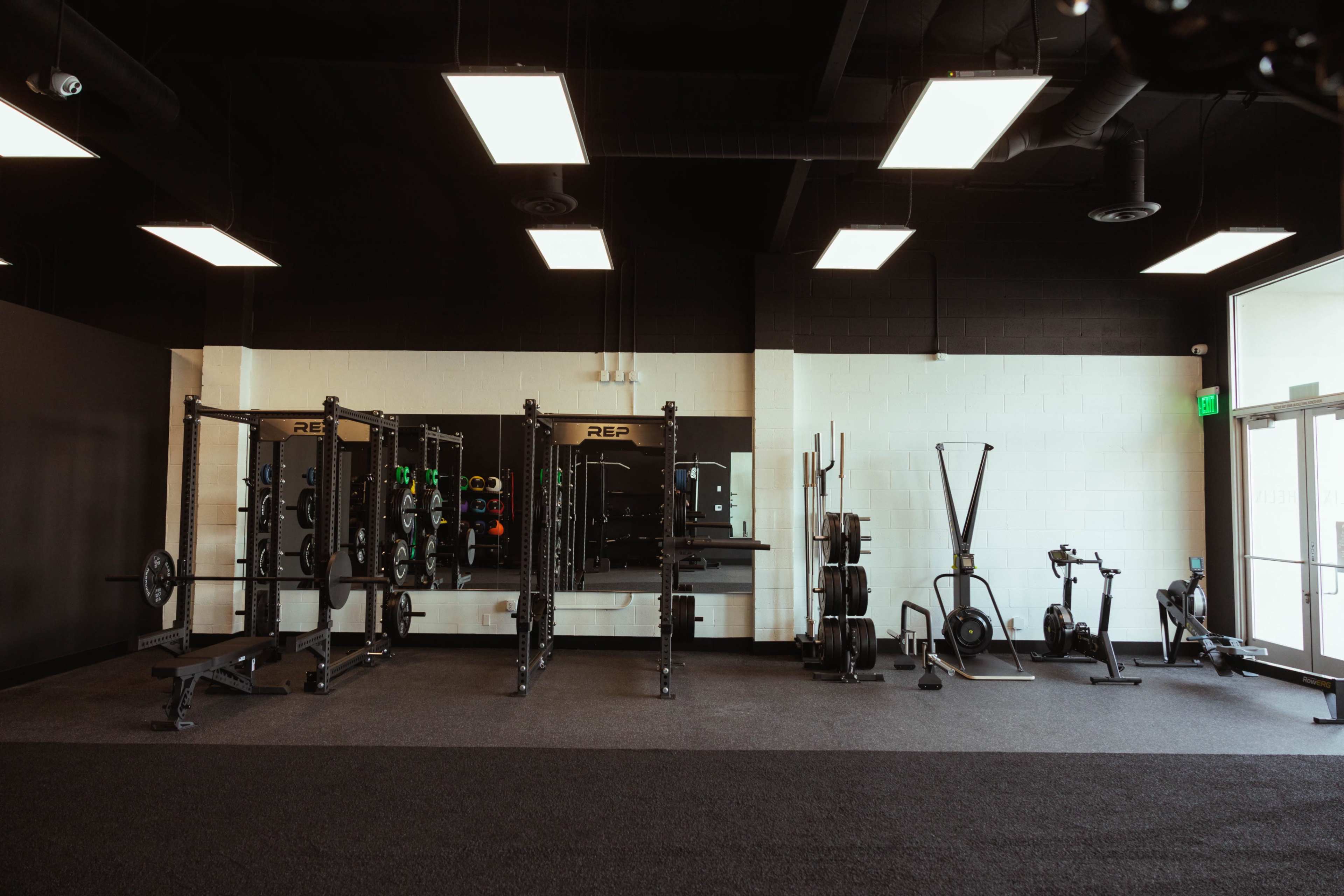 The image shows a spacious gym interior with weightlifting equipment, including squat racks and exercise machines, against a black and white backdrop.