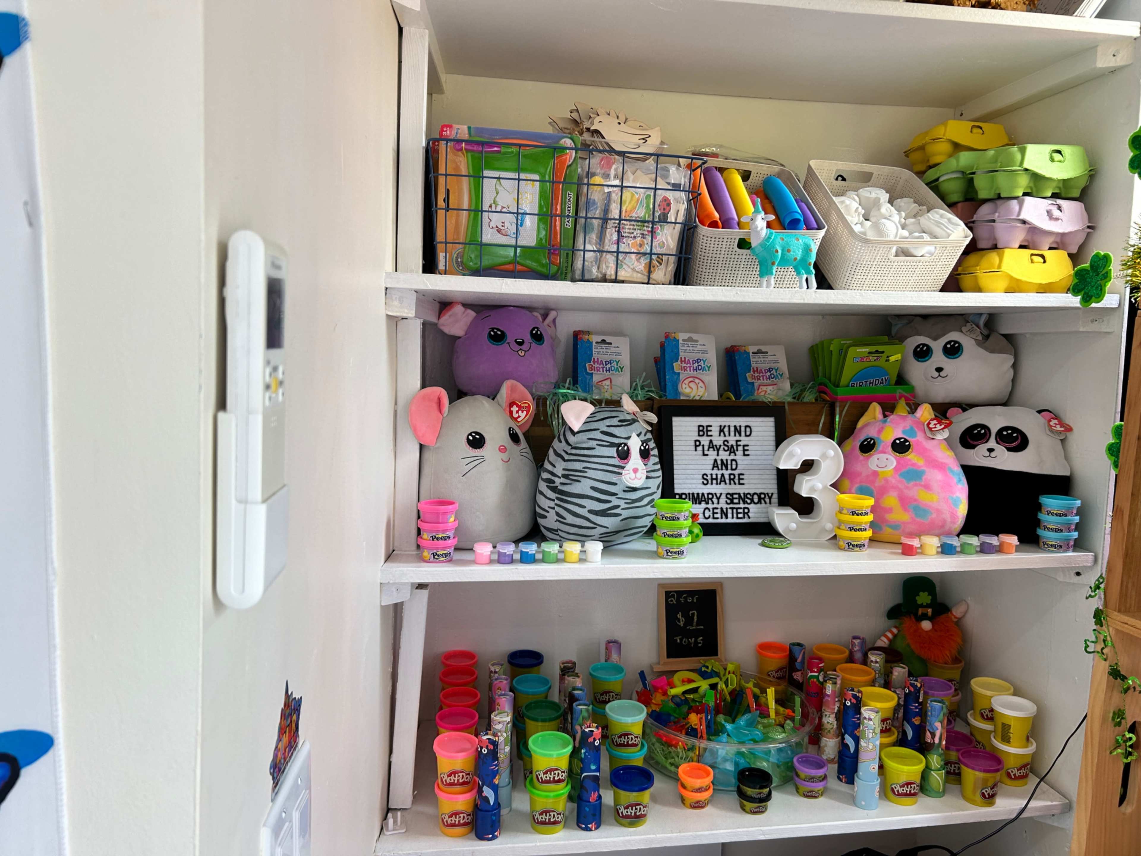 A shelving unit displays various colorful toys, art supplies, and plush animals, organized neatly alongside a sign that reads "BE KIND PLEASE AND SHARE."