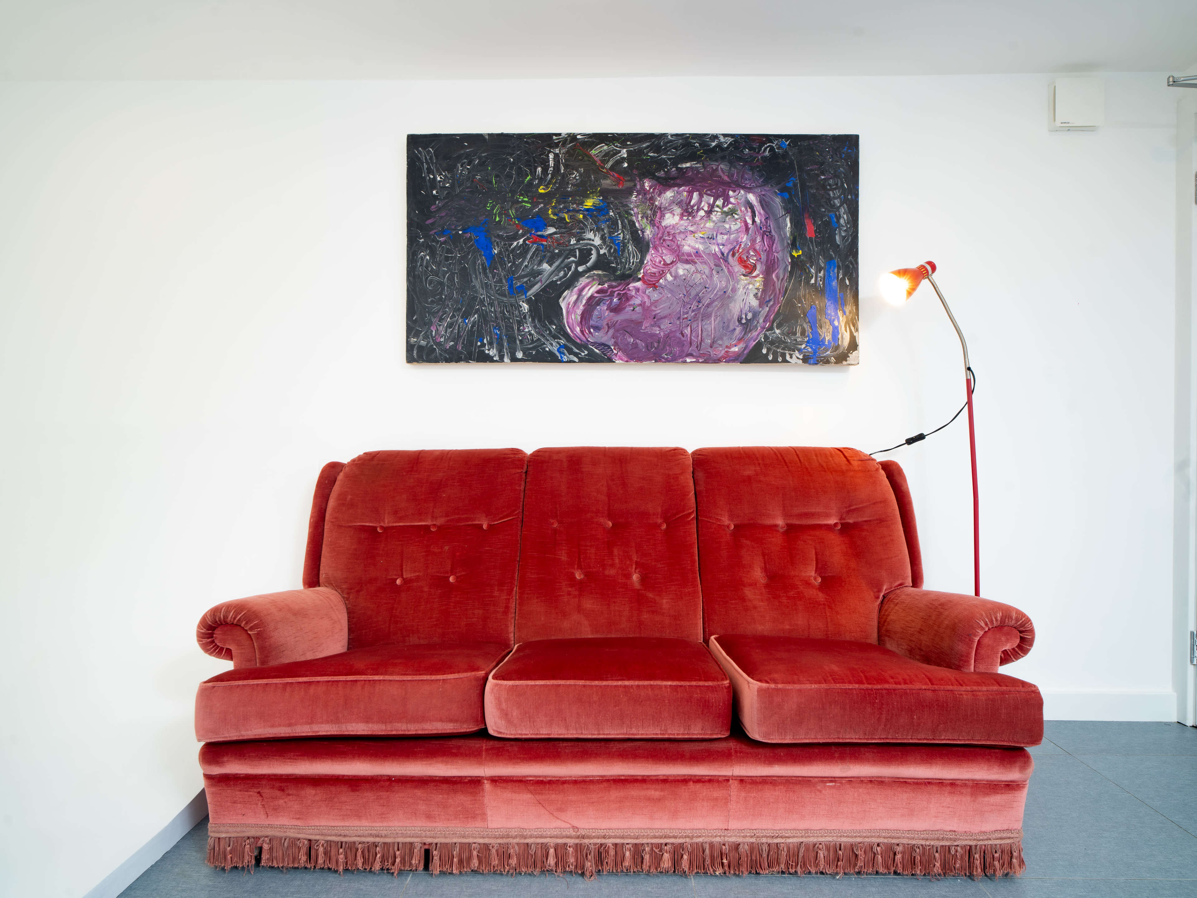 A red velvet sofa with a fringe sits against a white wall, beneath a colorful abstract painting and next to a floor lamp.