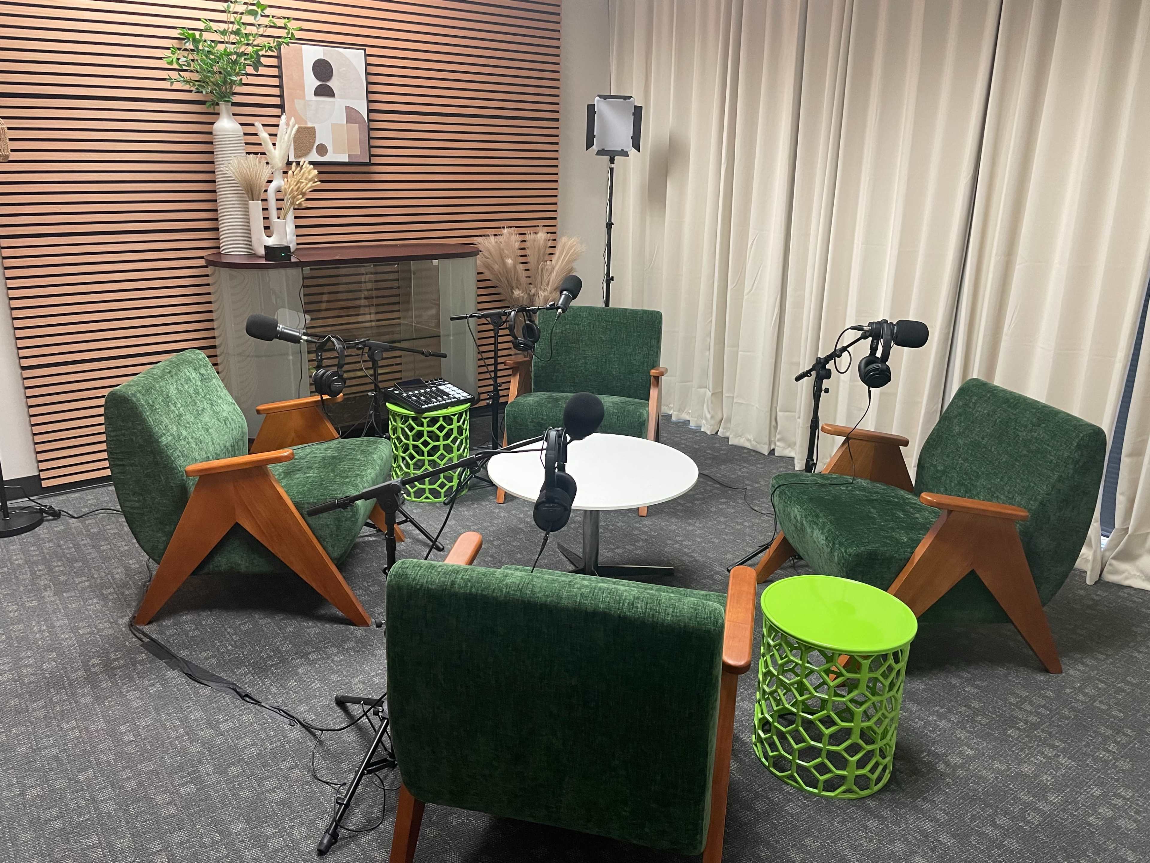 The image shows a modern discussion space with four green chairs arranged around a small white table, equipped with microphones and a decorative green side table.