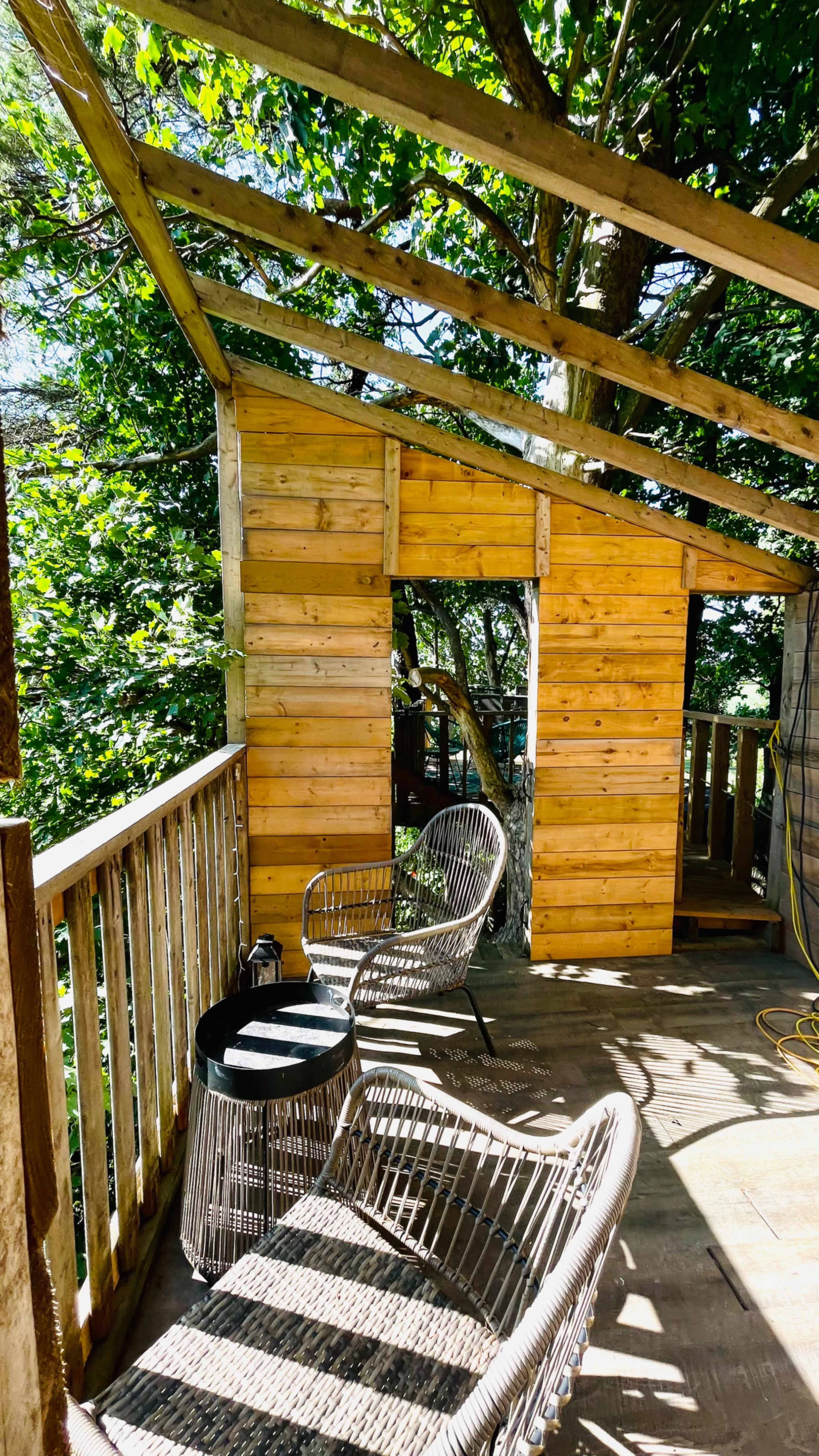 The image shows a wooden deck area with two chairs overlooking a partially constructed structure surrounded by trees.