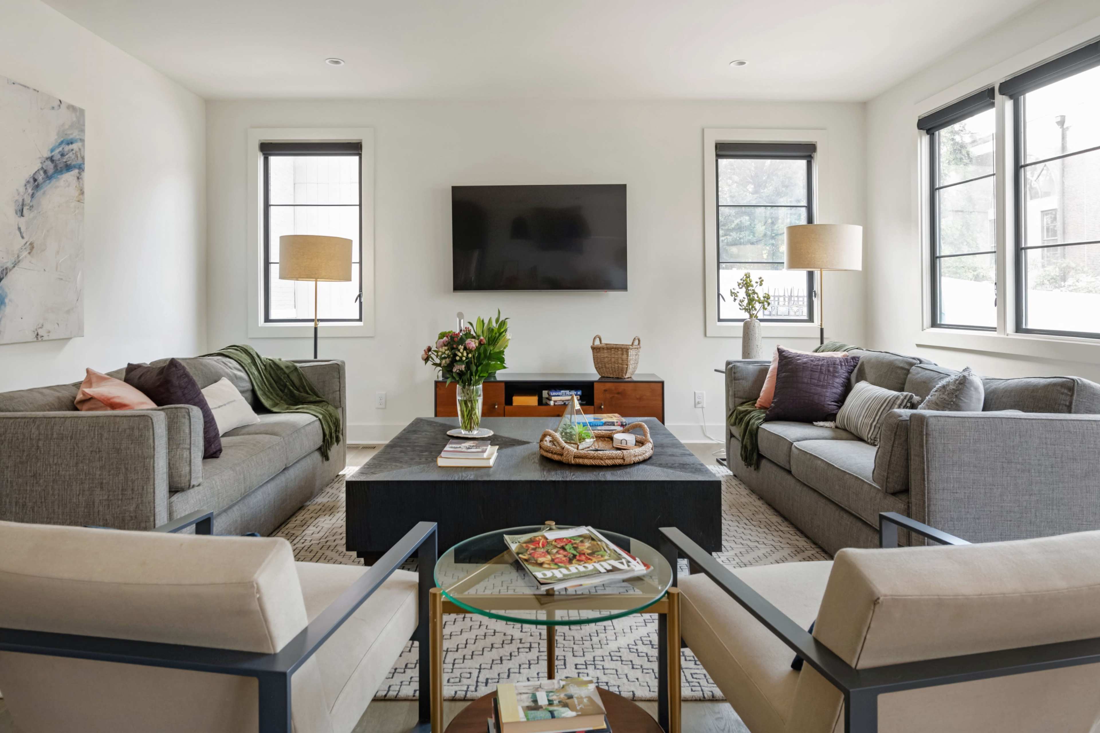 A bright living room features two gray sofas and two cream armchairs surrounding a coffee table, with a large television mounted on the wall and a floral arrangement on the table.