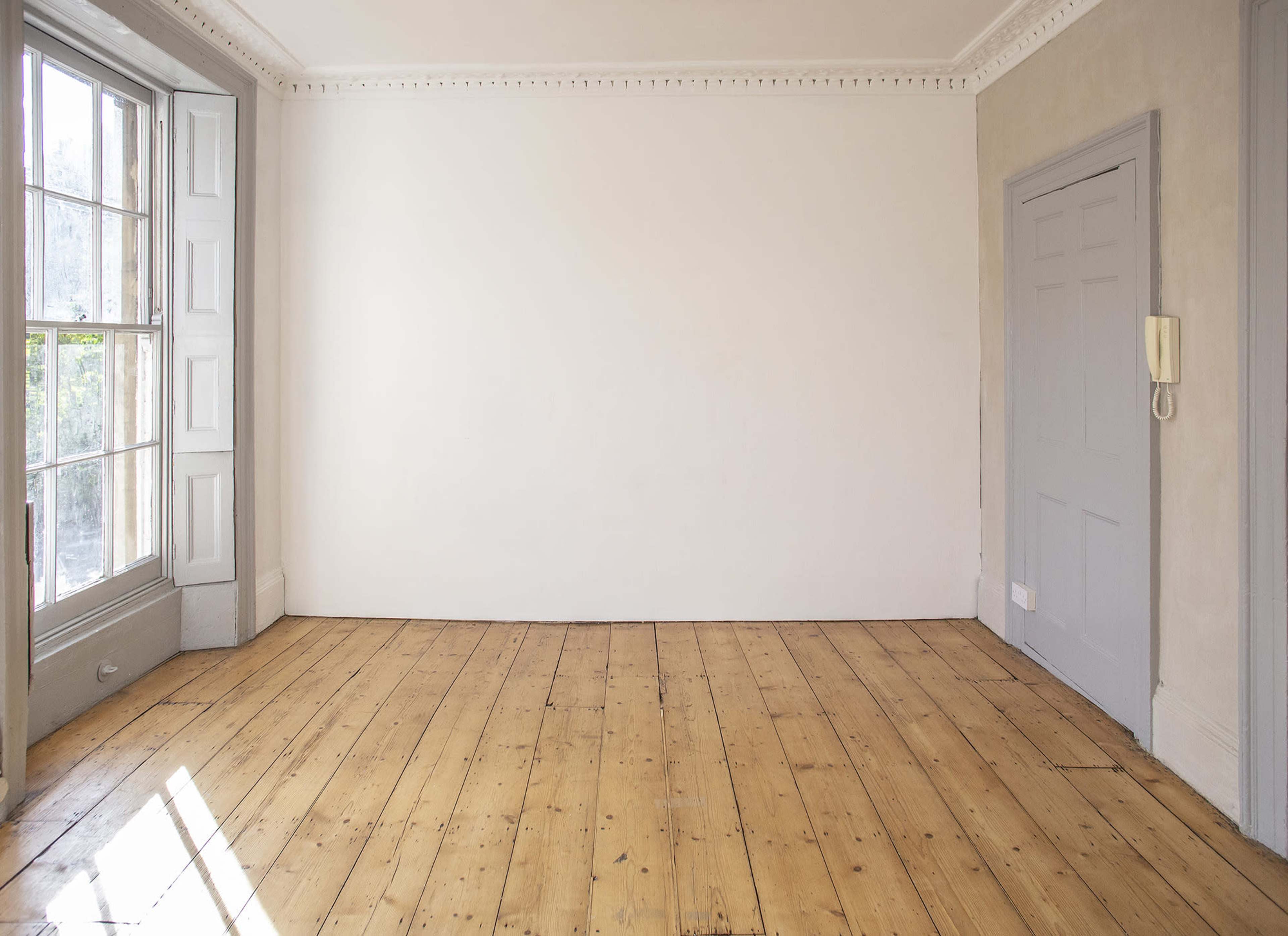 A well-lit, empty room features wooden flooring, a large window, and a plain wall with a door on one side.