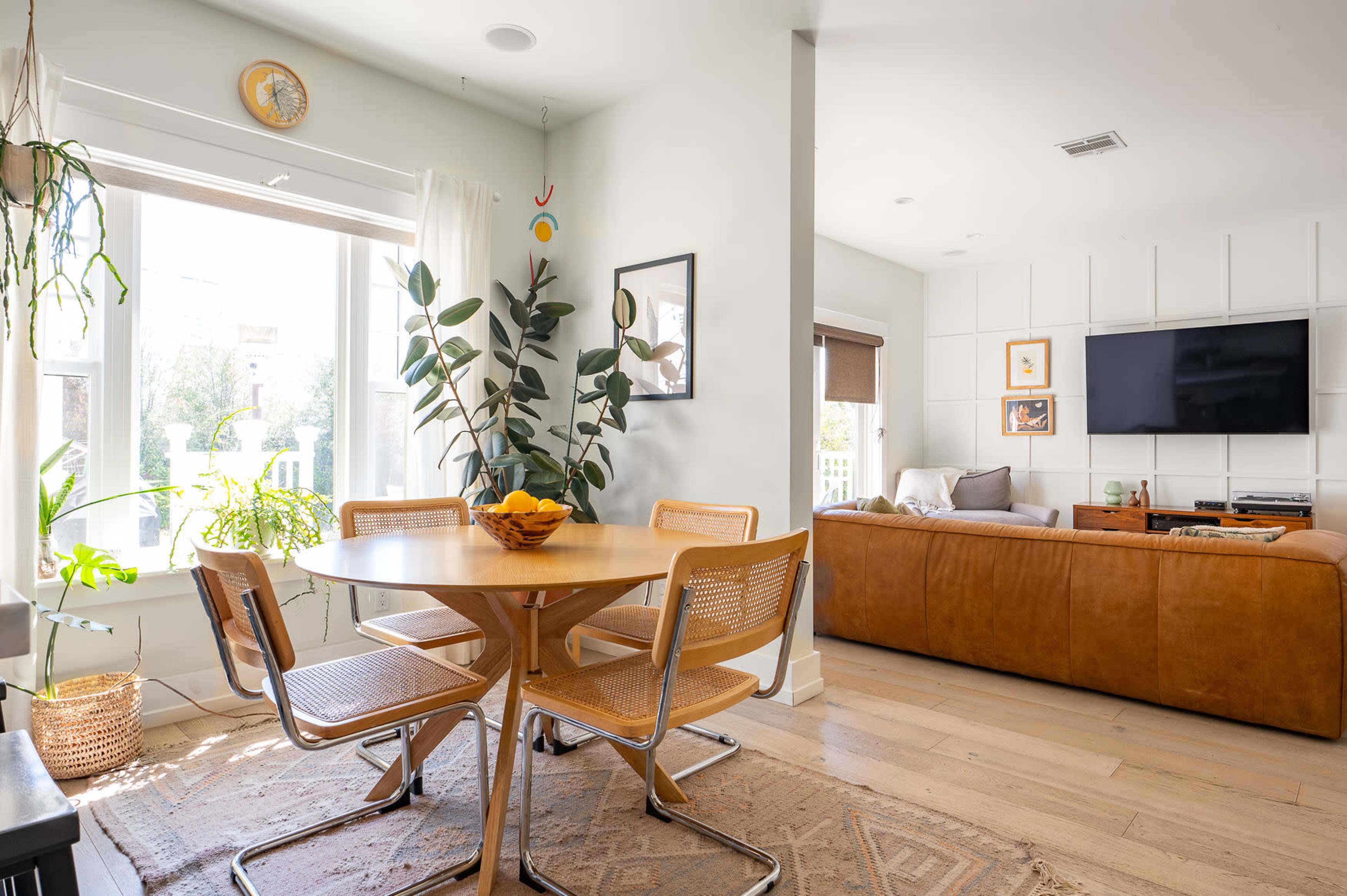 A cozy living space features a round dining table with four chairs and a sofa, surrounded by large windows and potted plants.