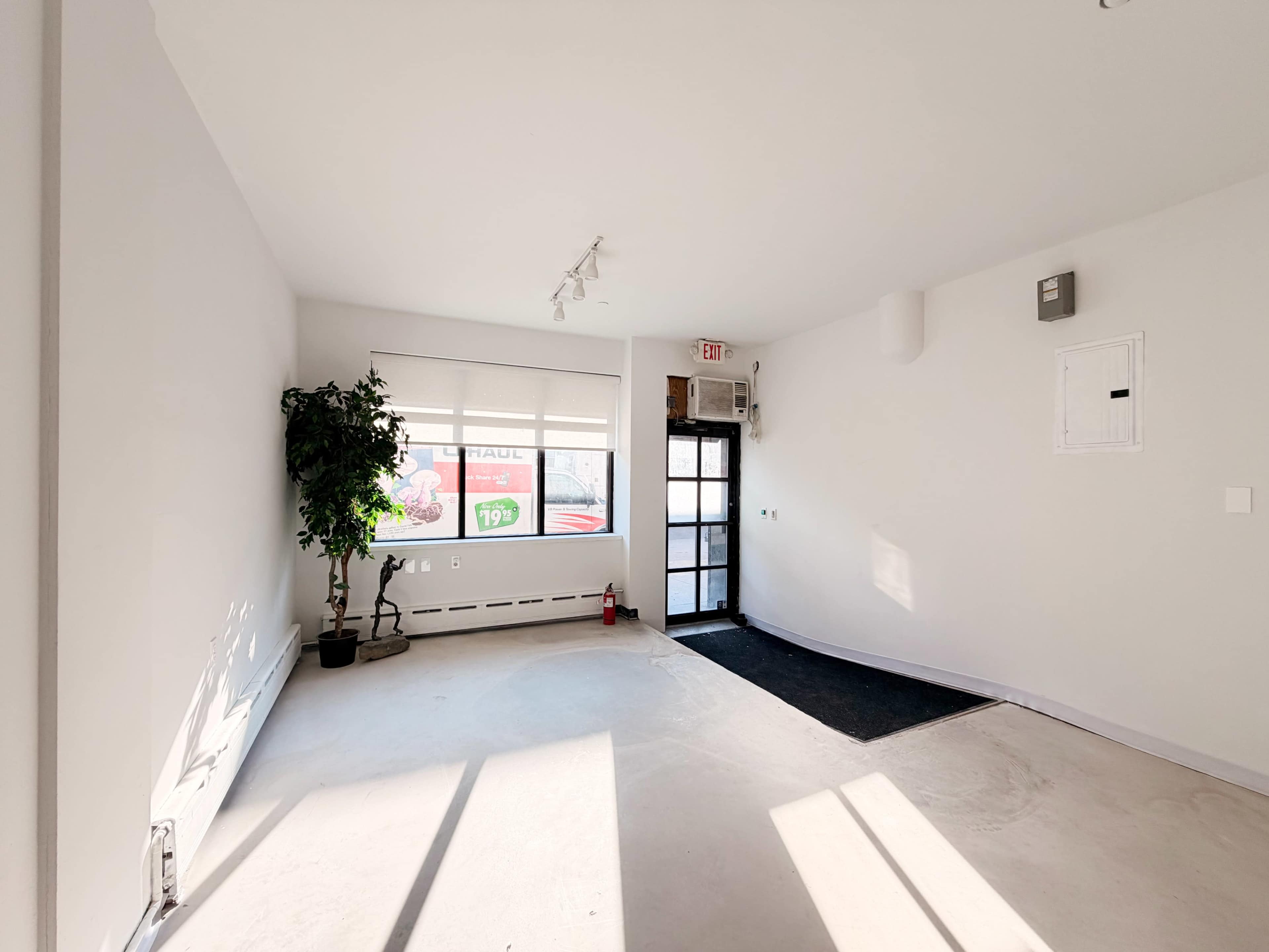 The image shows an empty, brightly lit room with a hardwood floor, a large window letting in natural light, and a doorway leading outside.