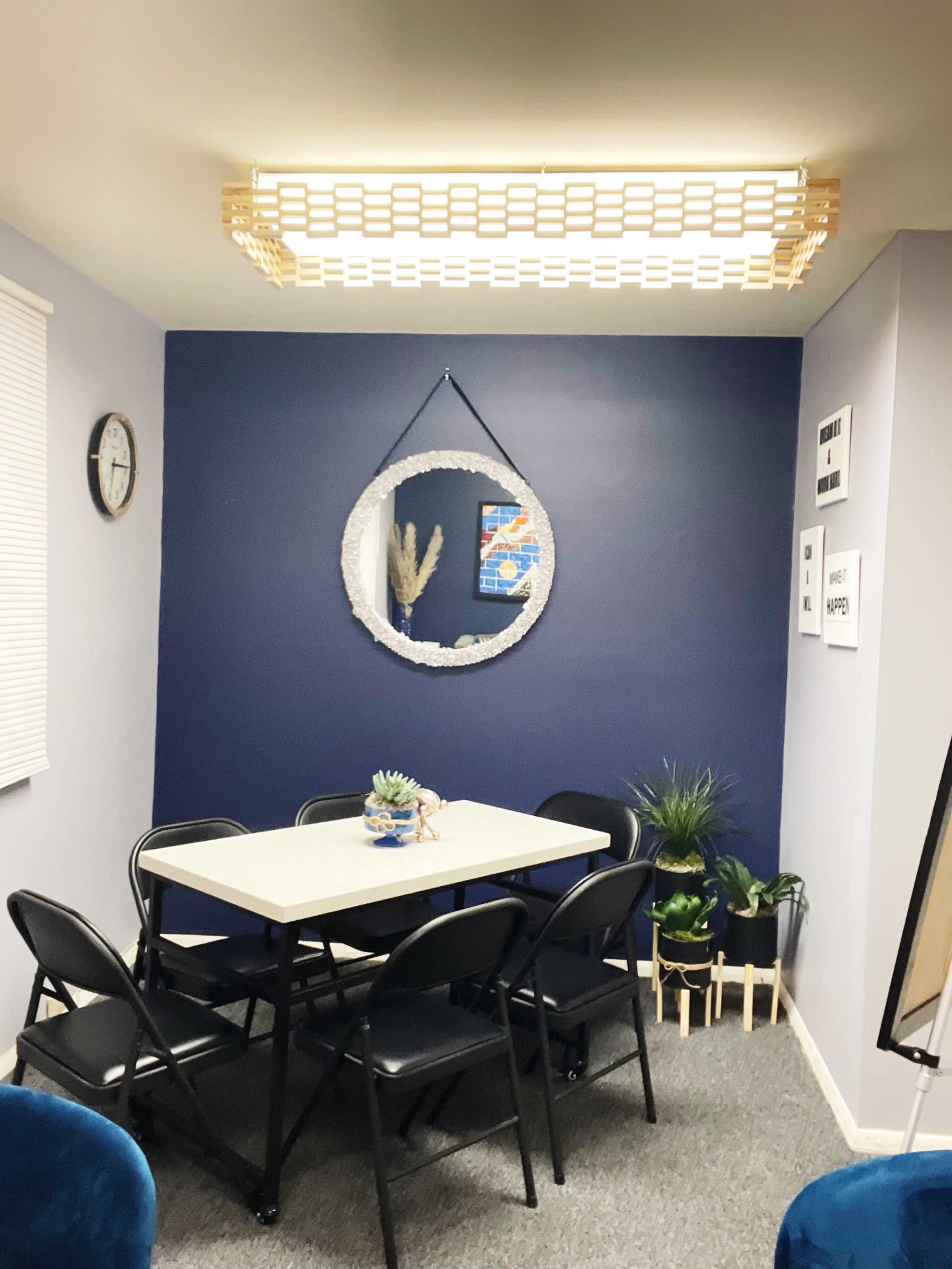 A small dining area with a round mirror on a dark blue wall, a white table surrounded by black chairs, and decorative plants in the corners.