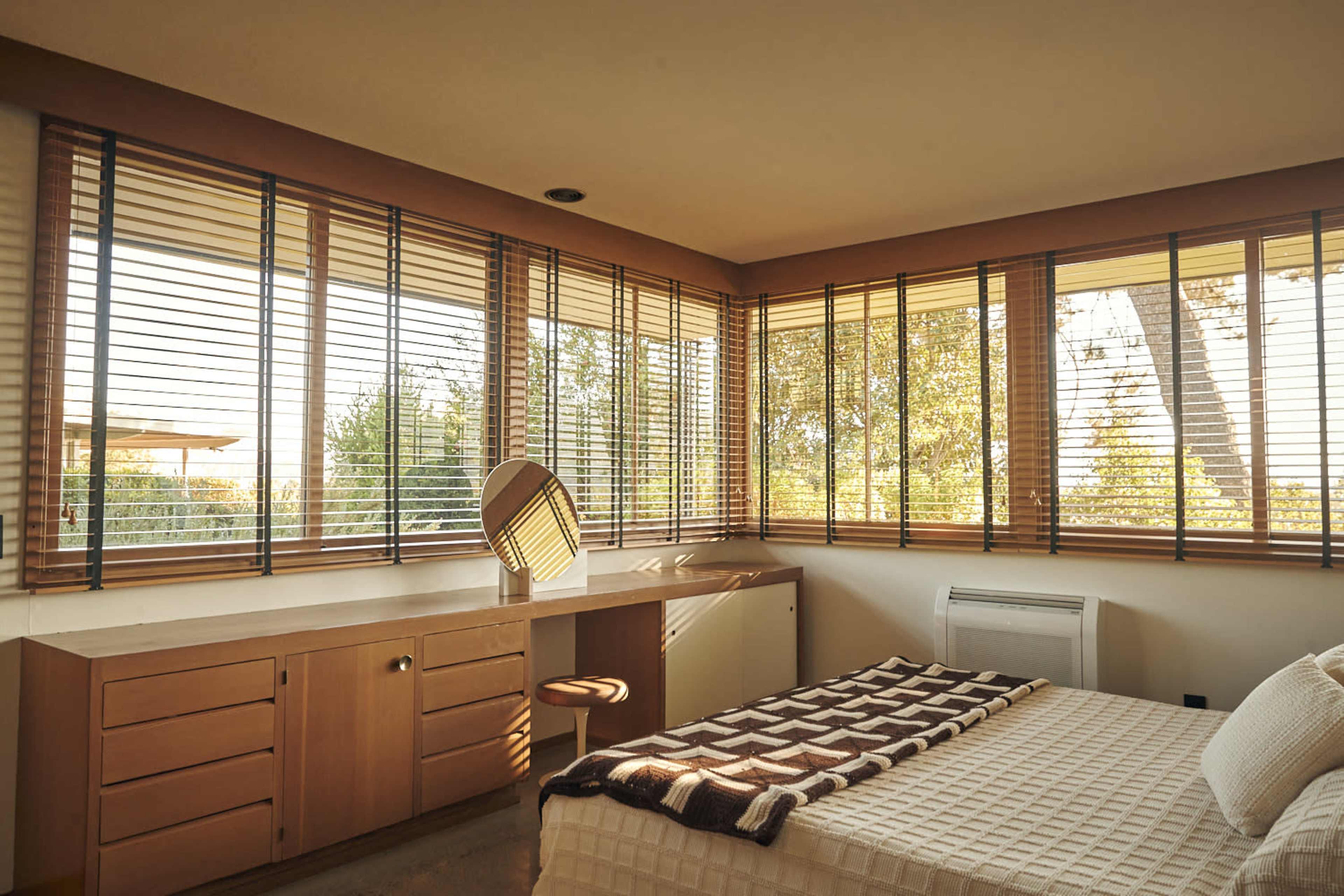 The image shows a spacious bedroom with large windows adorned with blinds, a bed with a textured blanket, and a vanity mirror on a wooden desk.