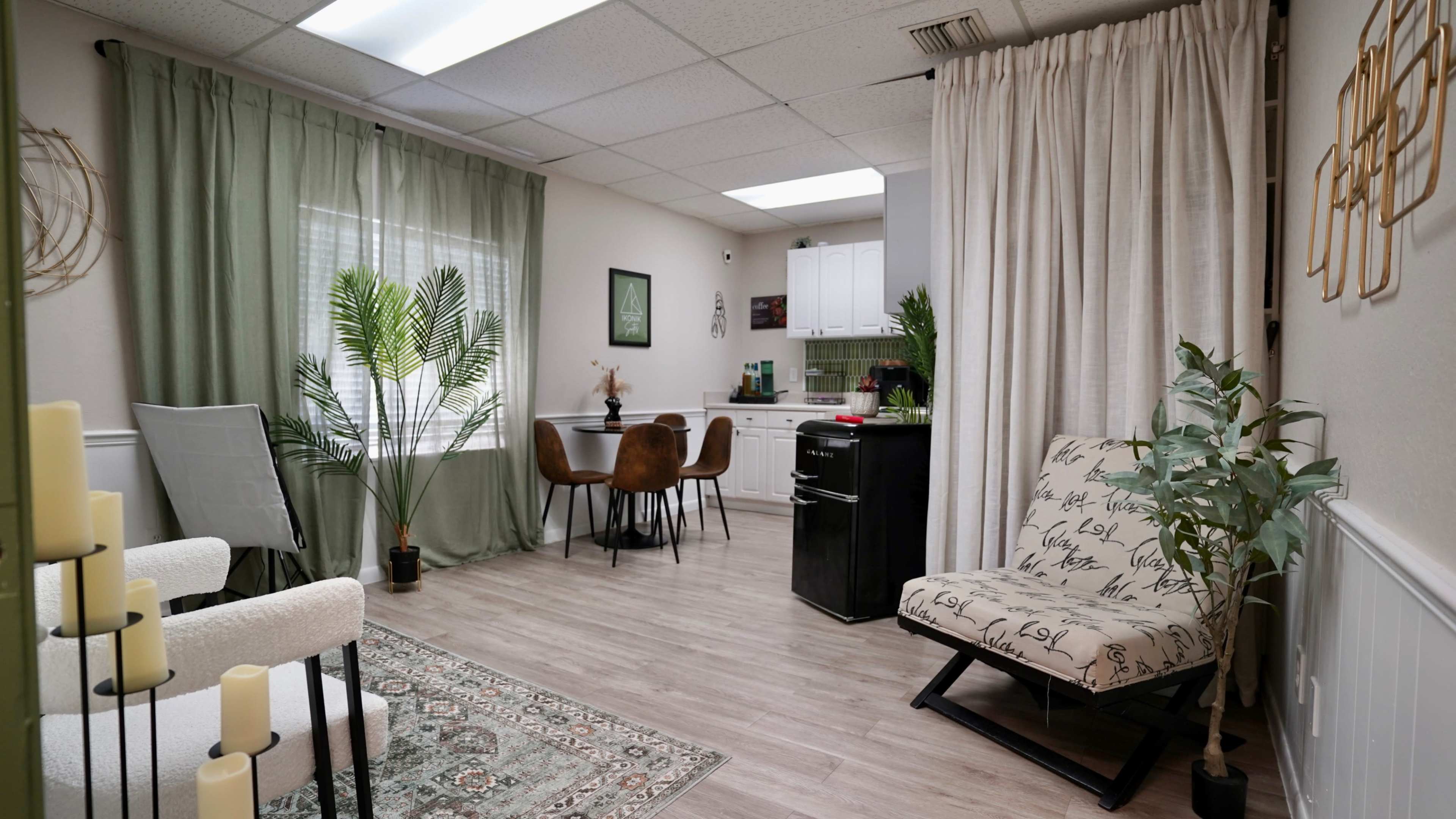 The image shows a modern, well-lit room featuring a seating area with a patterned chair, a small dining table, and a kitchenette with white cabinets, light flooring, and decorative plants.