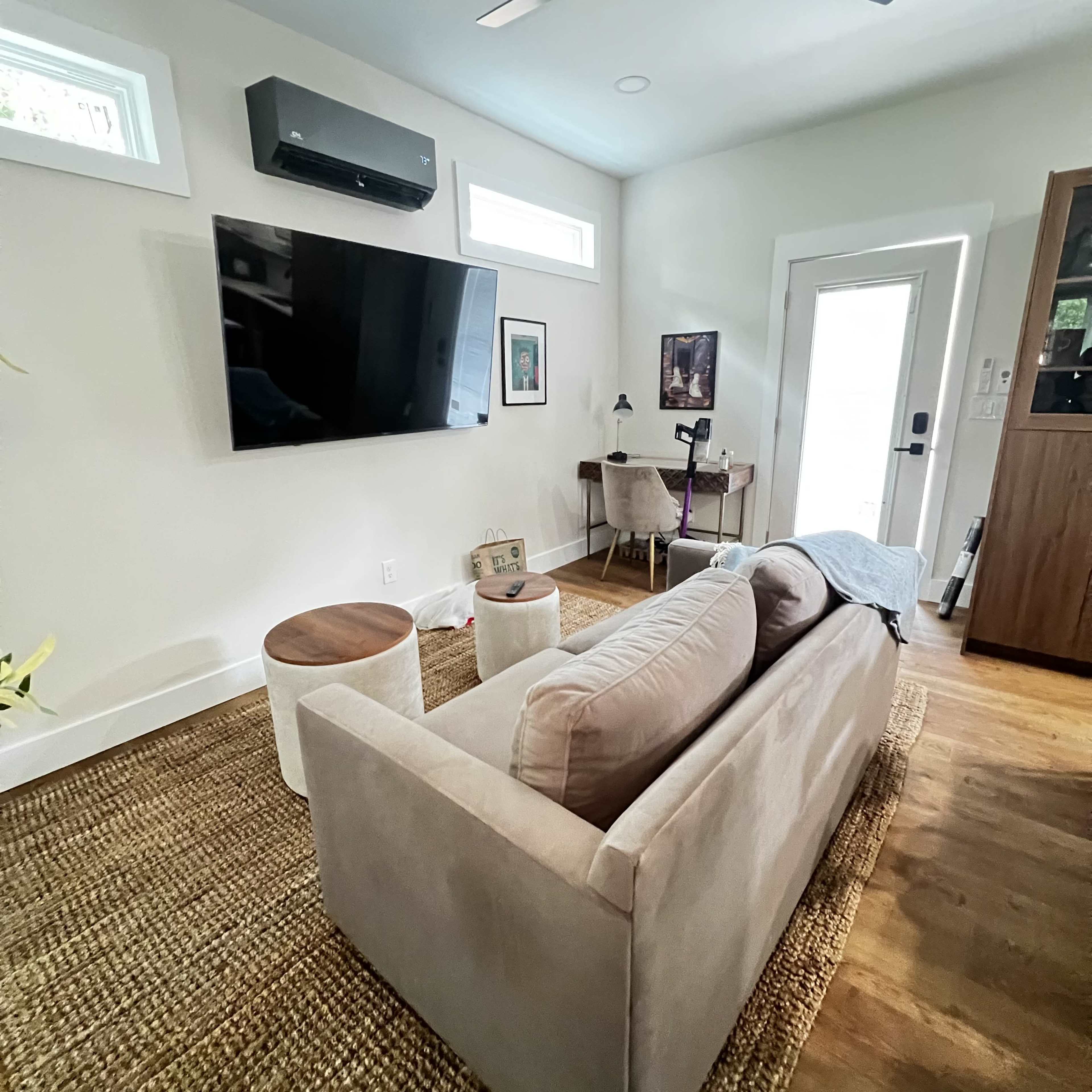 A living room features a sofa facing a mounted television, with a small table and workspace near a doorway.