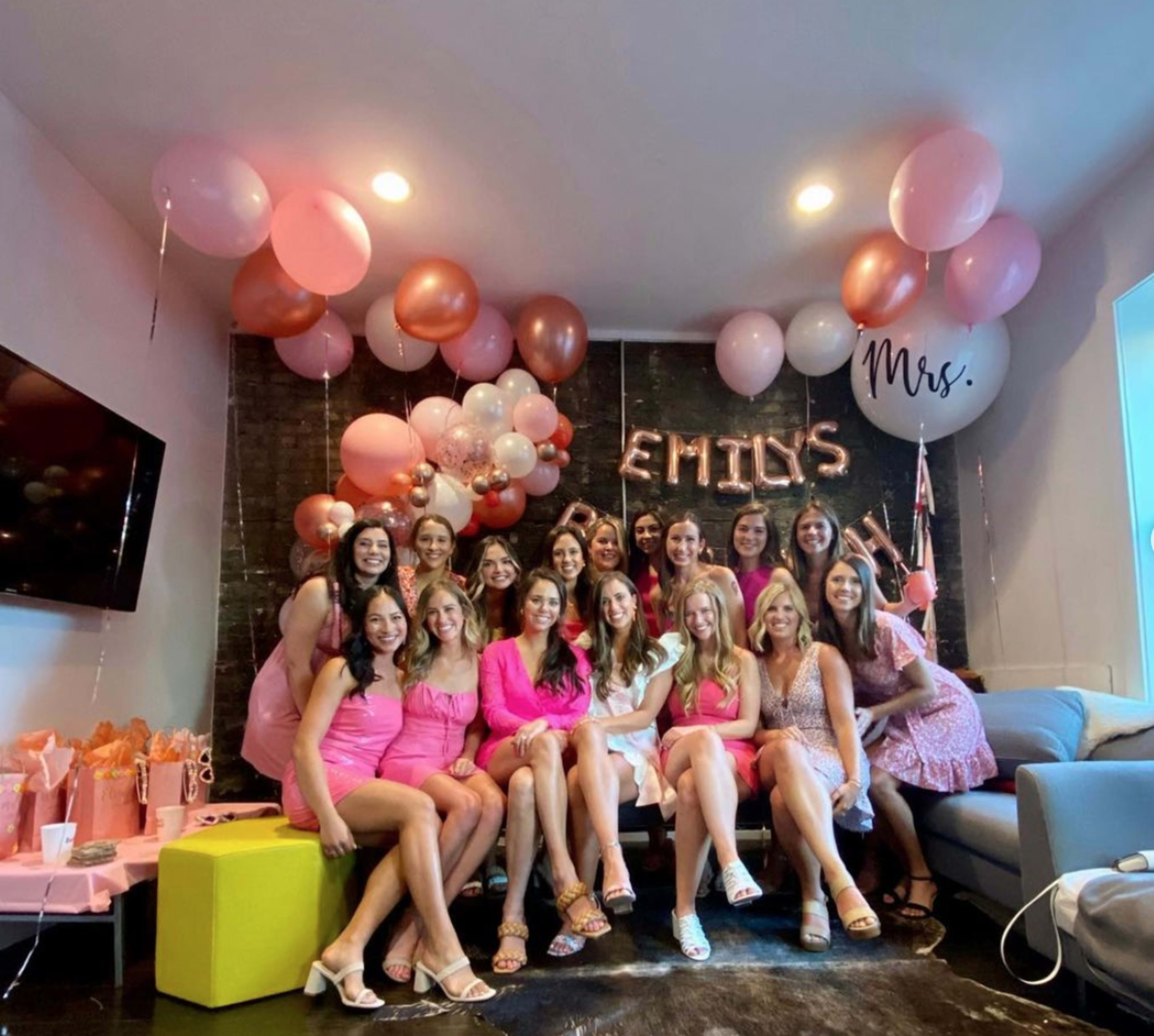 A large group of women dressed in pink attire gather in a decorated room with balloons and a sign that says "Mrs." for a celebration.