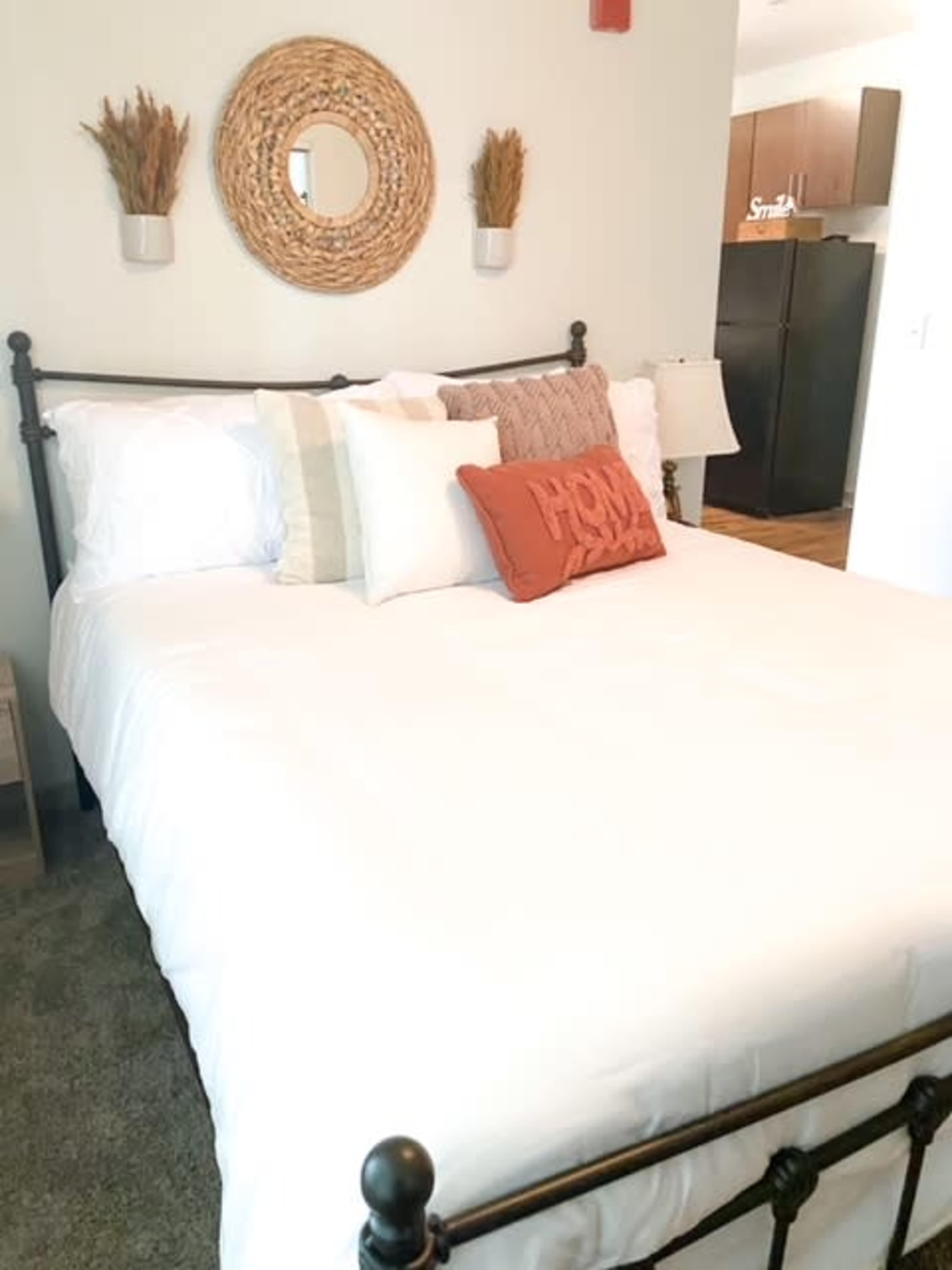 The image shows a neatly made bed with decorative pillows, a round mirror above it, and a nightstand beside the bed in a well-lit room.