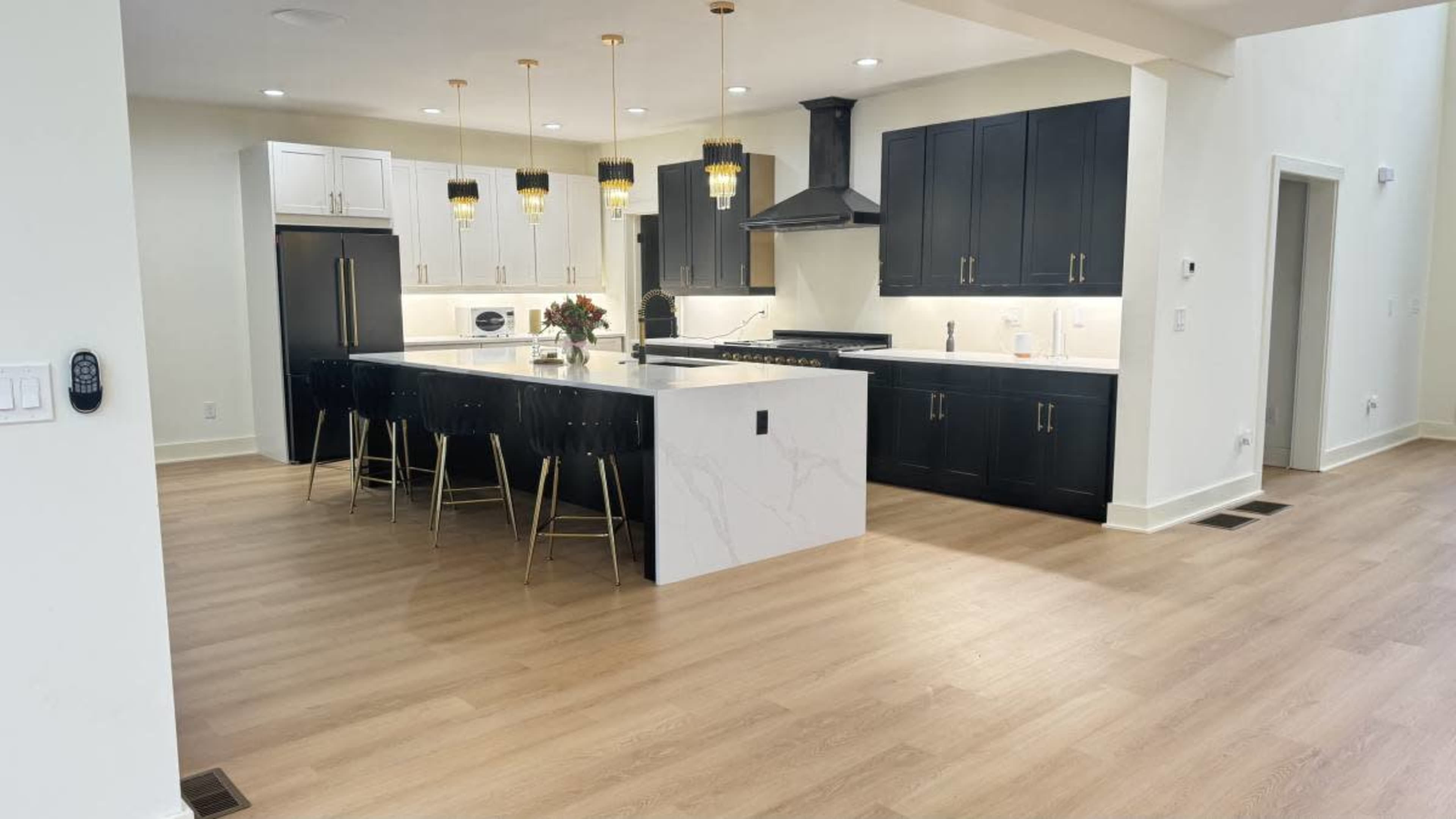 The image shows a modern kitchen featuring a large central island with a white marble surface, black cabinetry, gold light fixtures, and stainless steel appliances.