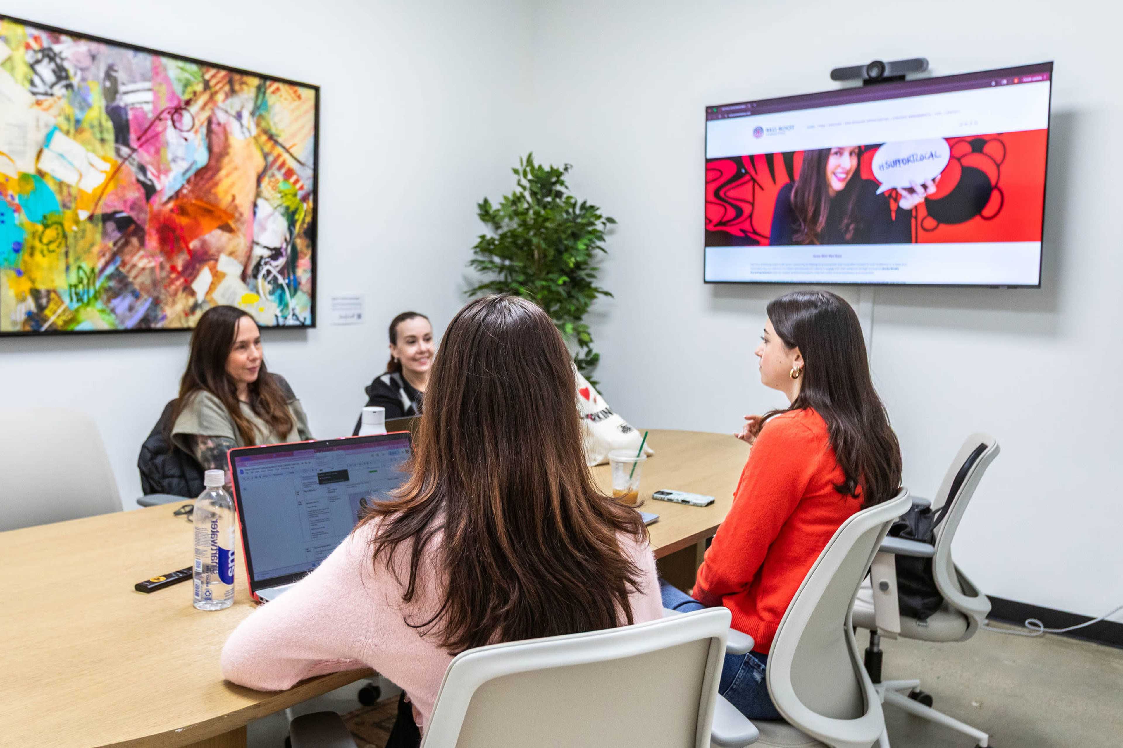 A group of three women sits around a conference table in a meeting room, with a large screen displaying a website in the background.