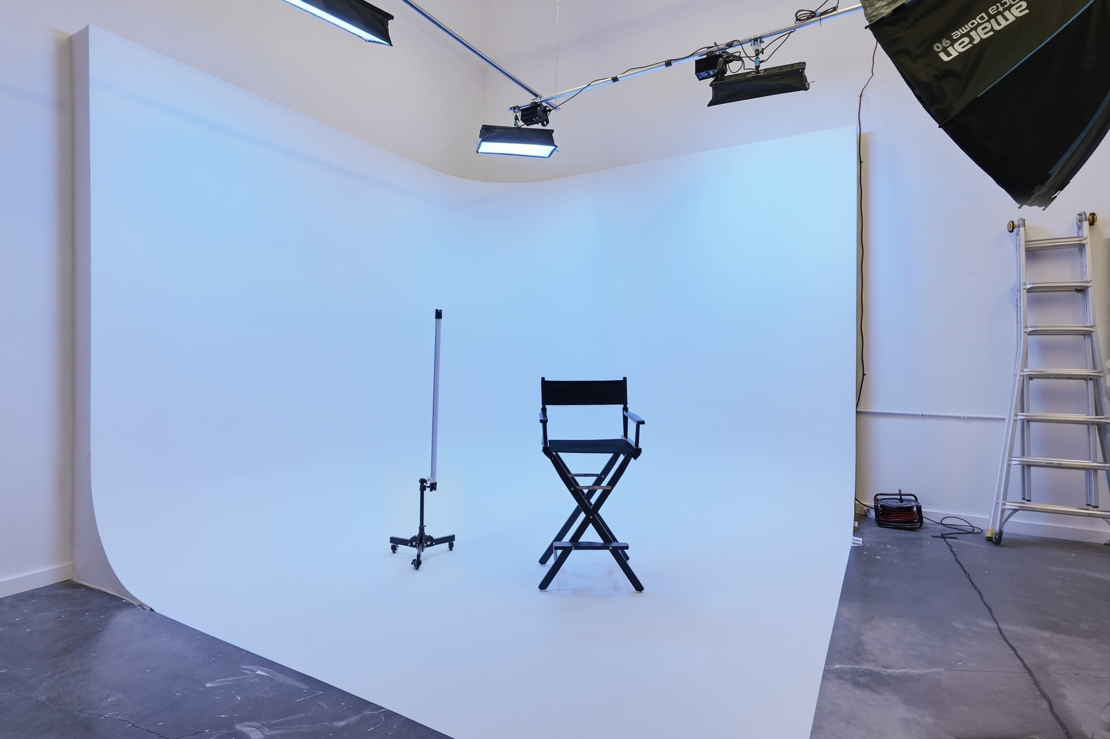 The image shows a photography studio with a empty white backdrop, a black director's chair, and several overhead lights.