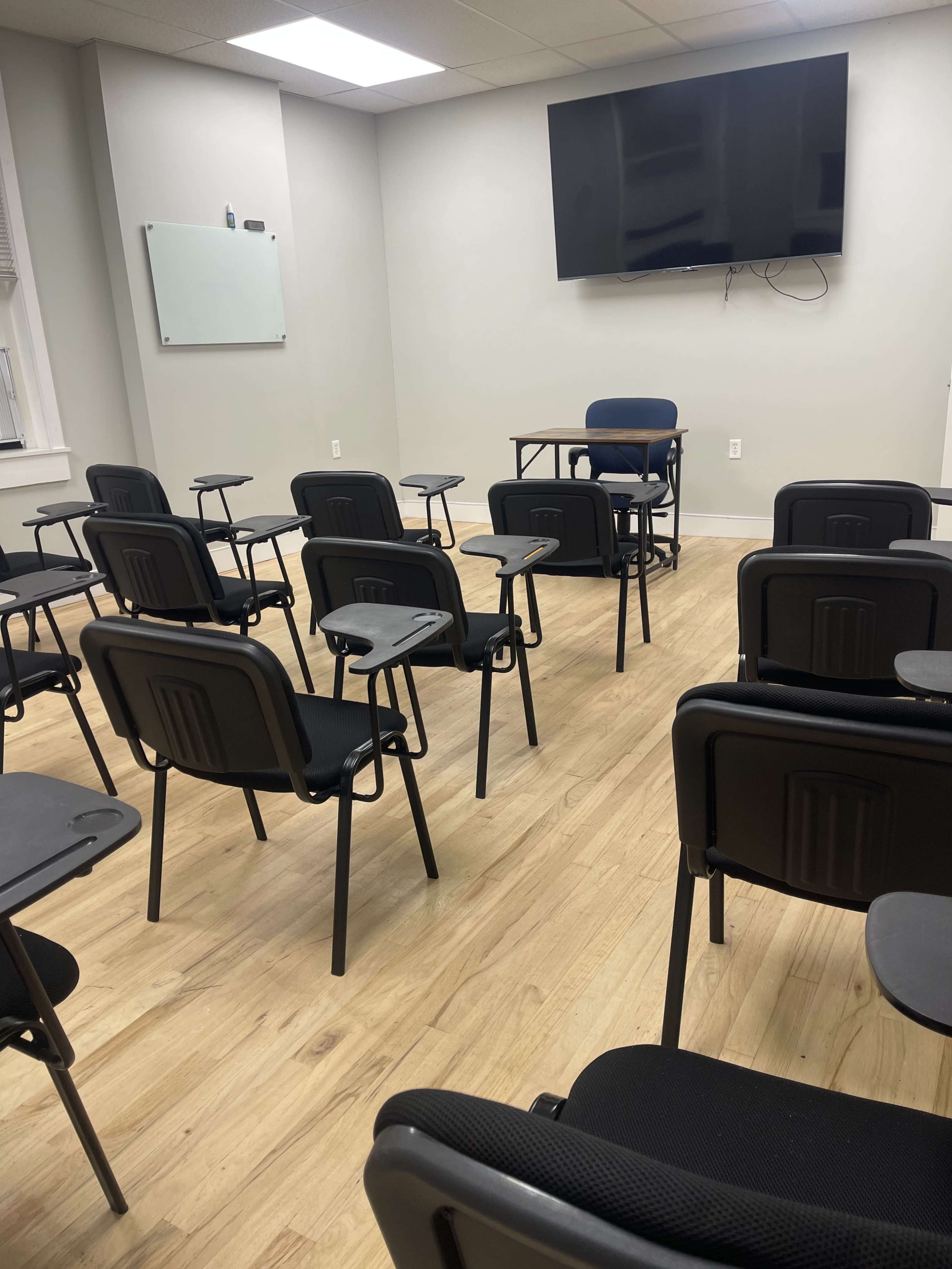 The image shows a small classroom with rows of black chairs arranged facing a front area that features a television and a single chair beside a table.