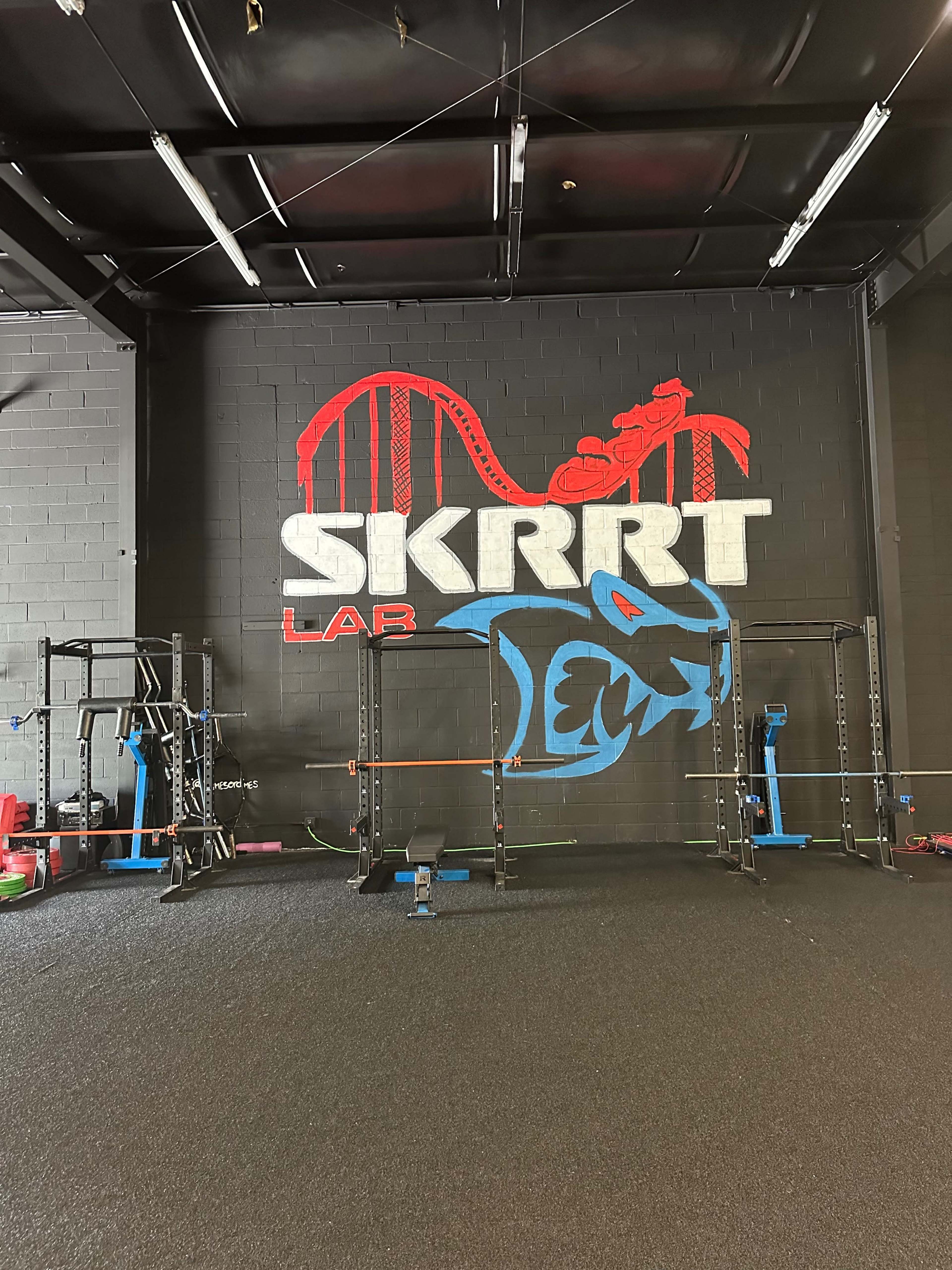 A gym interior featuring a large mural with the text "SKRRT" and a blue graphic of a dragon, alongside various gym equipment on the floor.