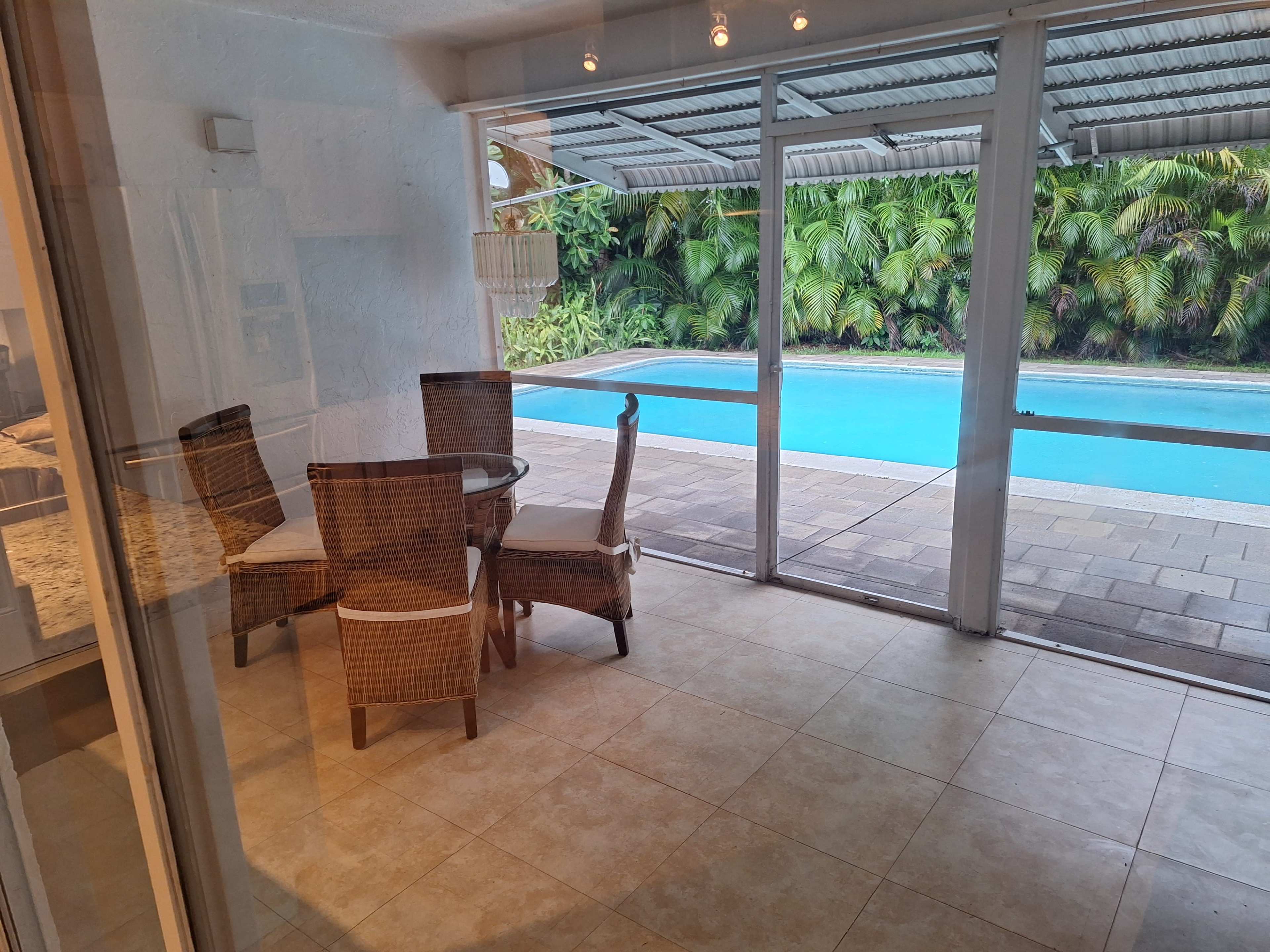 A glass-enclosed room features a dining table with four wicker chairs, overlooking a swimming pool surrounded by tropical foliage.