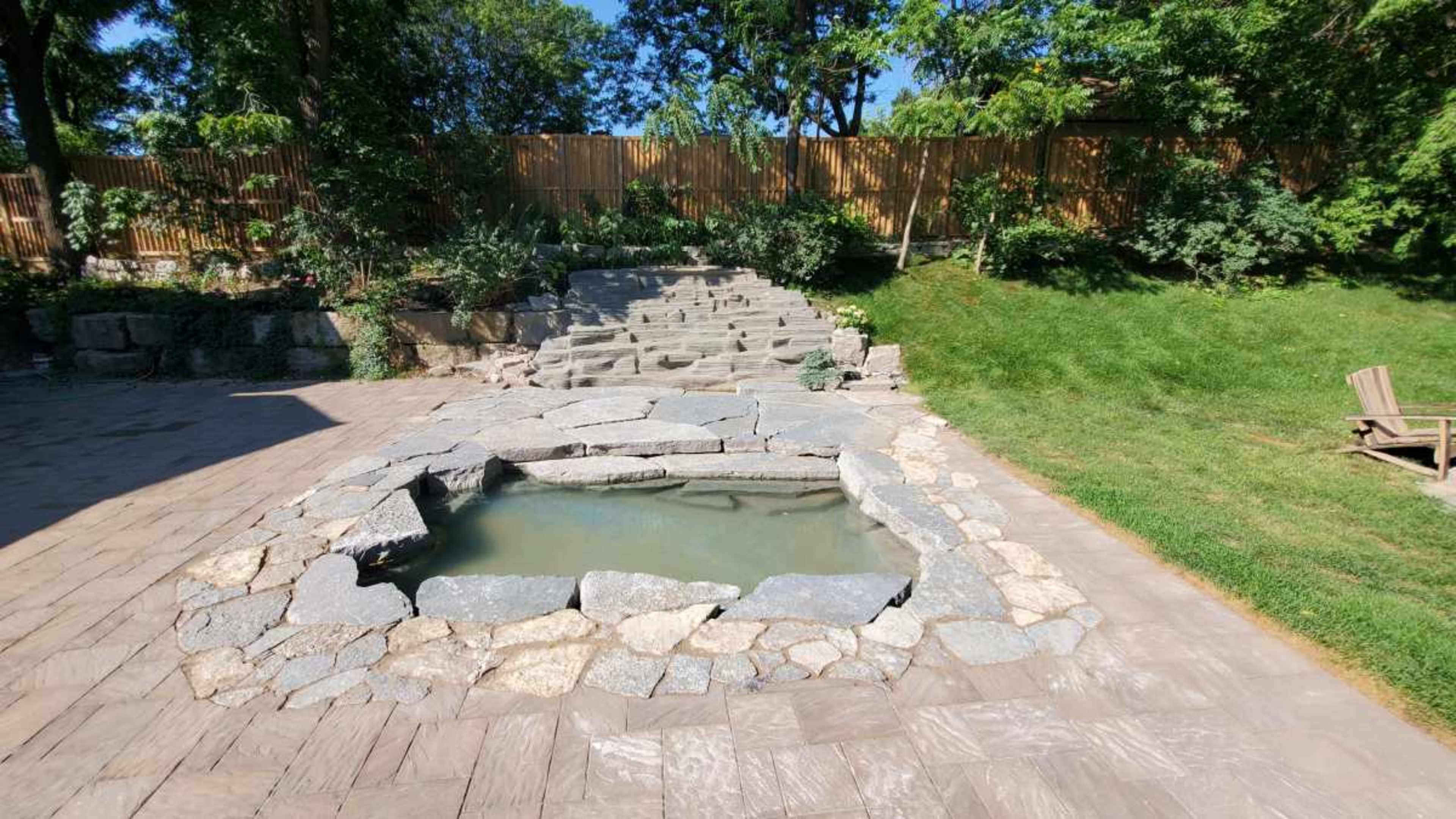 A stone-lined, shallow pool is situated at the bottom of a set of stone steps in a landscaped yard.