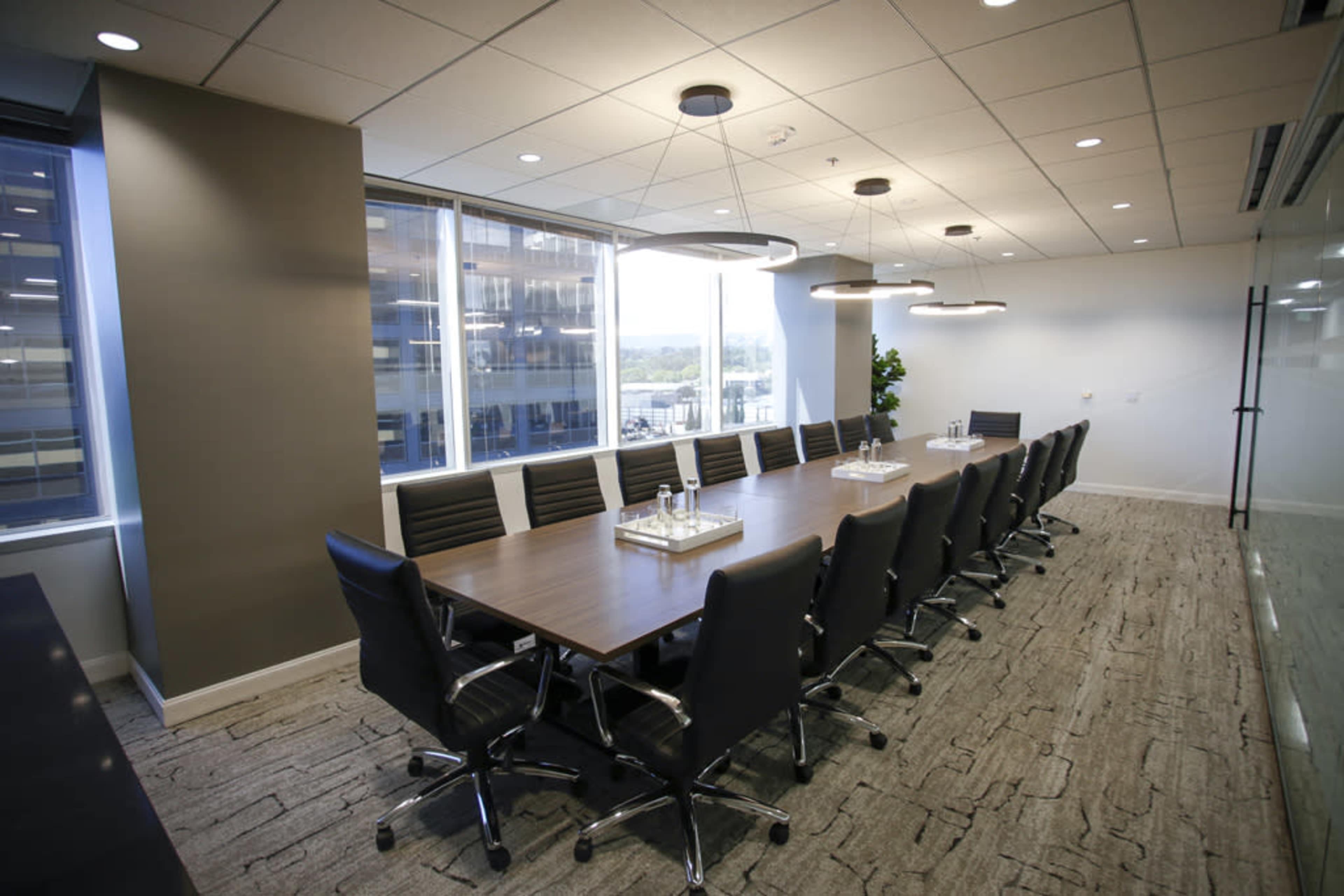 A large conference room features a long wooden table surrounded by black chairs, with windows providing natural light and a view of the outdoors.