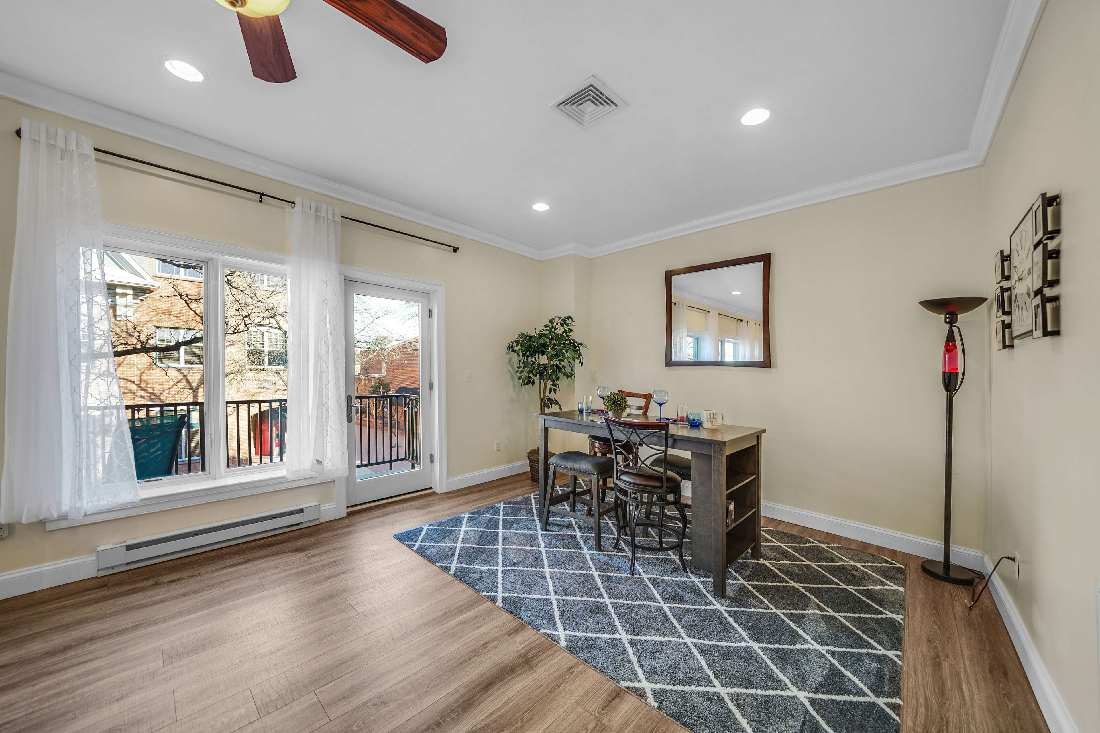 A bright dining area features a wooden table with chairs, large windows overlooking a balcony, and light-colored walls with a geometric area rug.