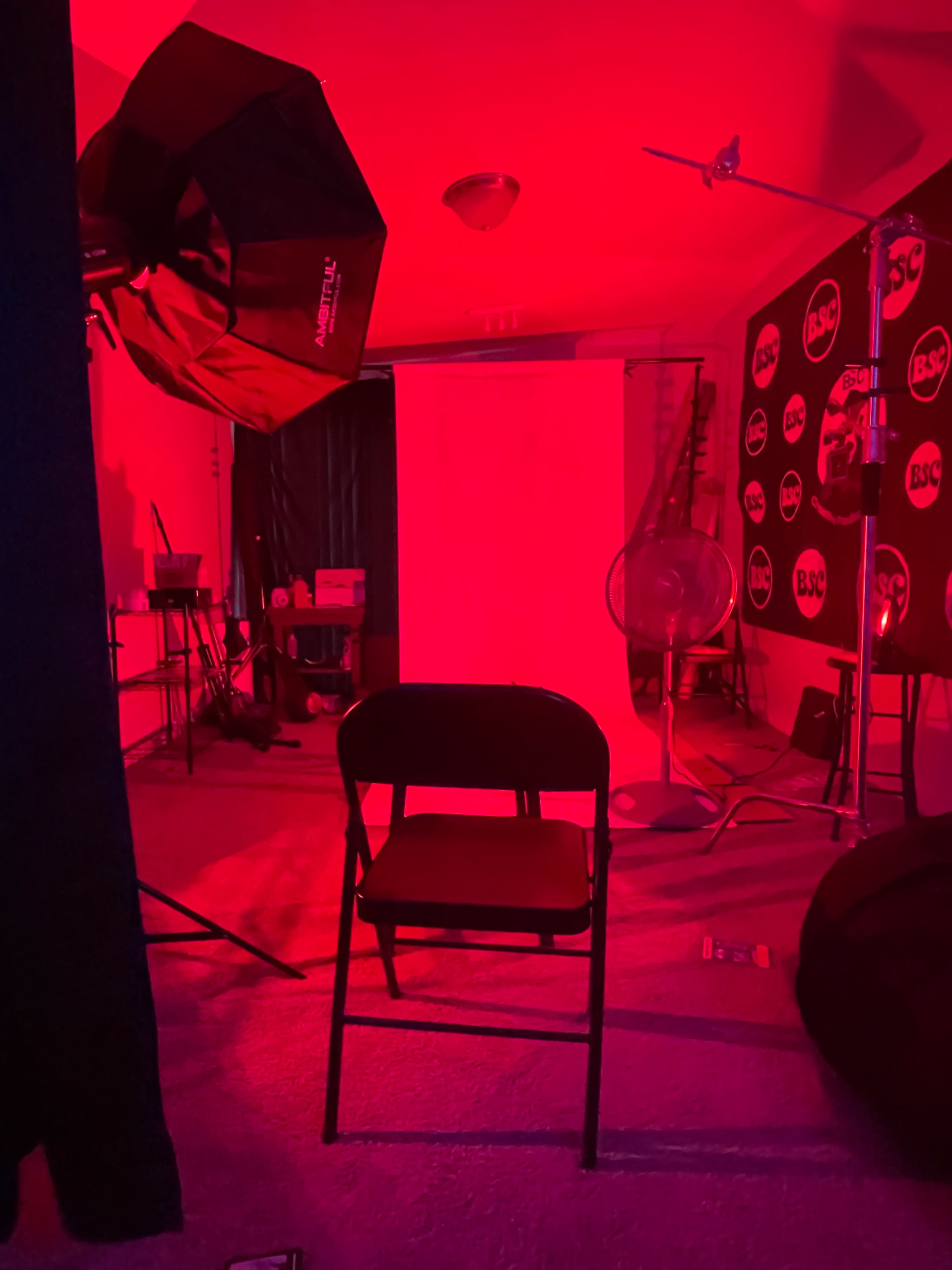 A room with a red-lit backdrop, a black folding chair, and studio lighting equipment.