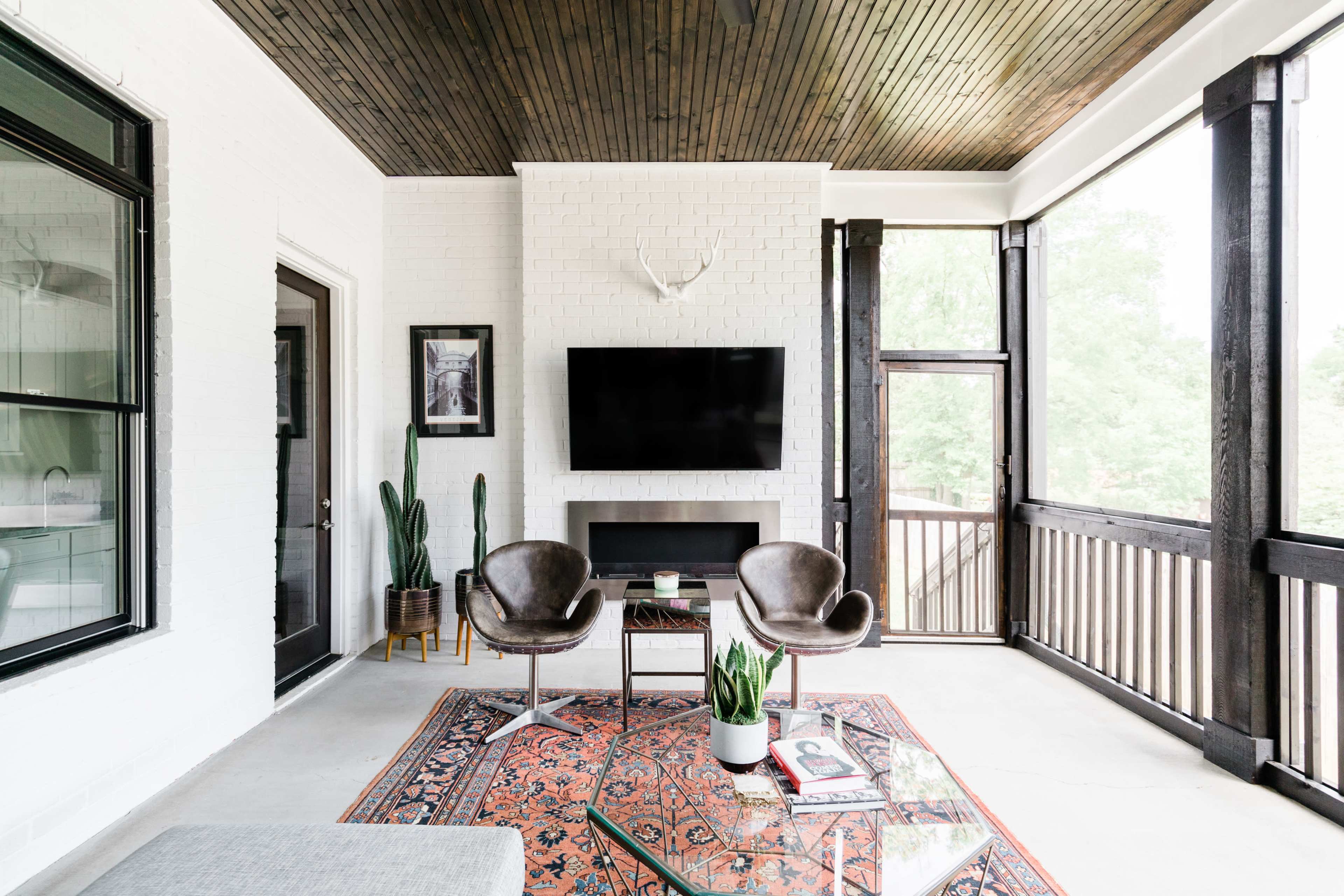 The image shows a screened-in porch featuring two dark chairs, a coffee table, a television mounted on a white brick wall, and plants, with wooden accents and a patterned rug.