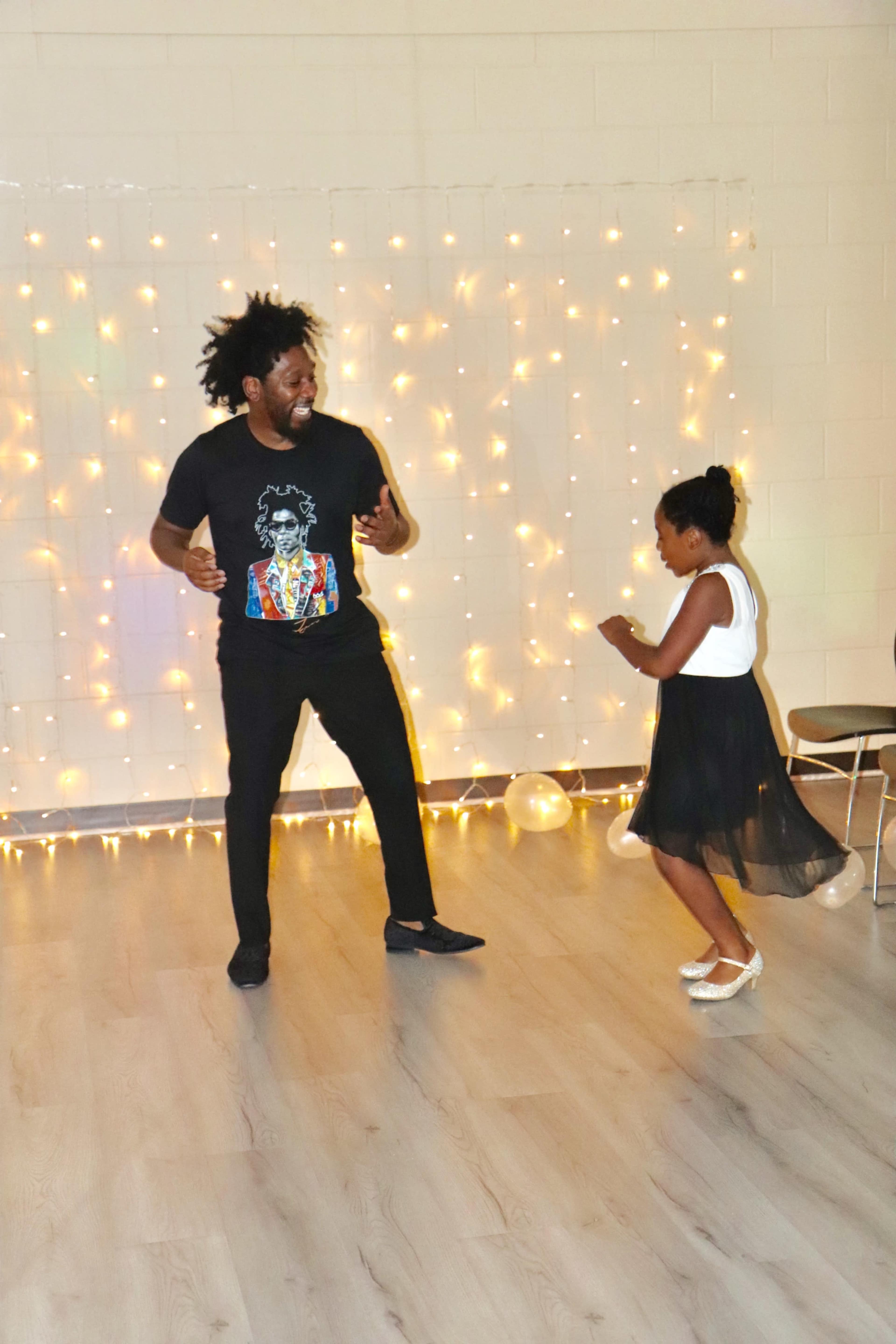 A man and a girl are dancing joyfully together in front of a backdrop of twinkling string lights.
