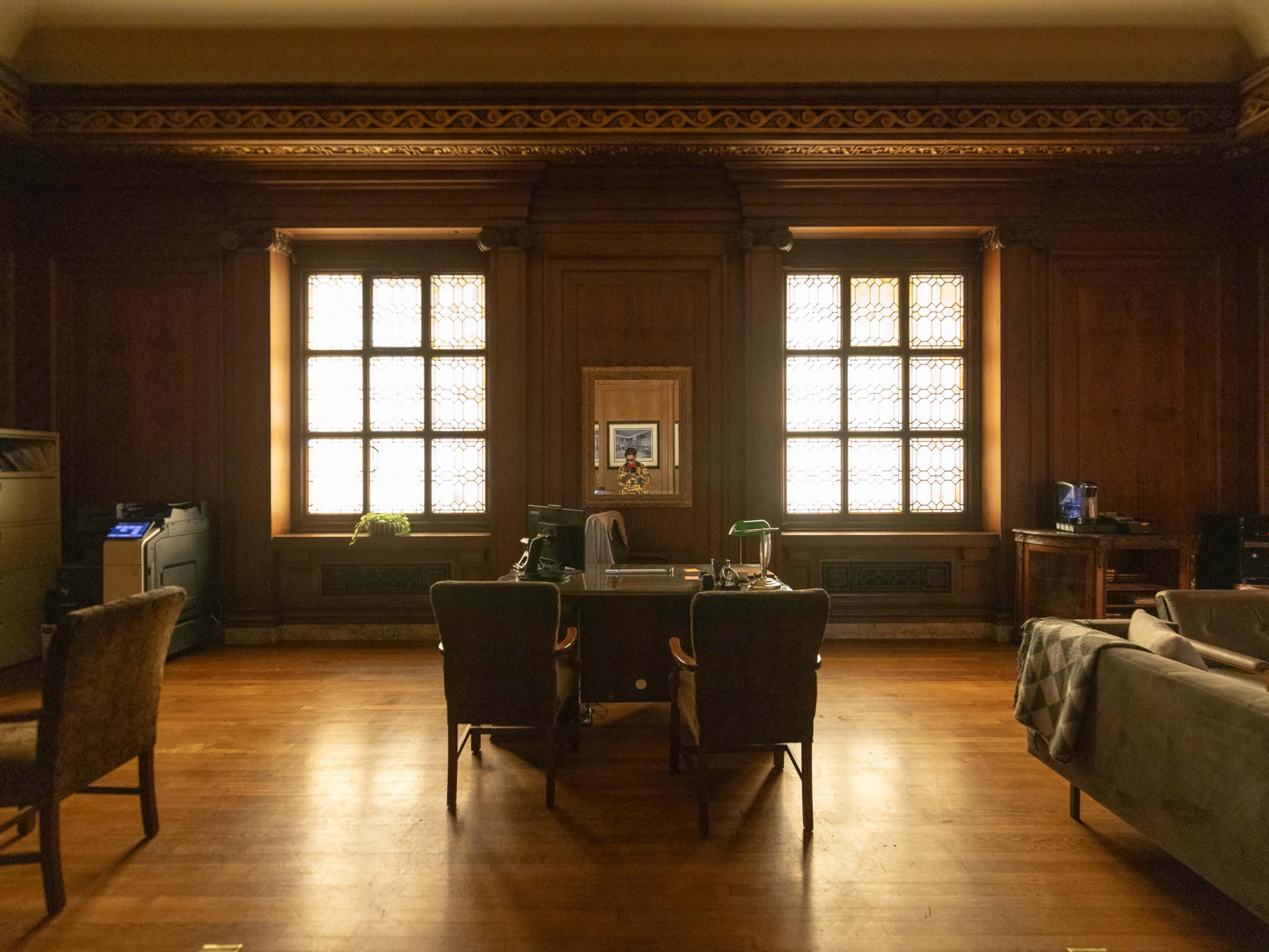 The image shows a dimly lit room with wooden paneling, two chairs facing a desk, and large windows adorned with patterned glass.