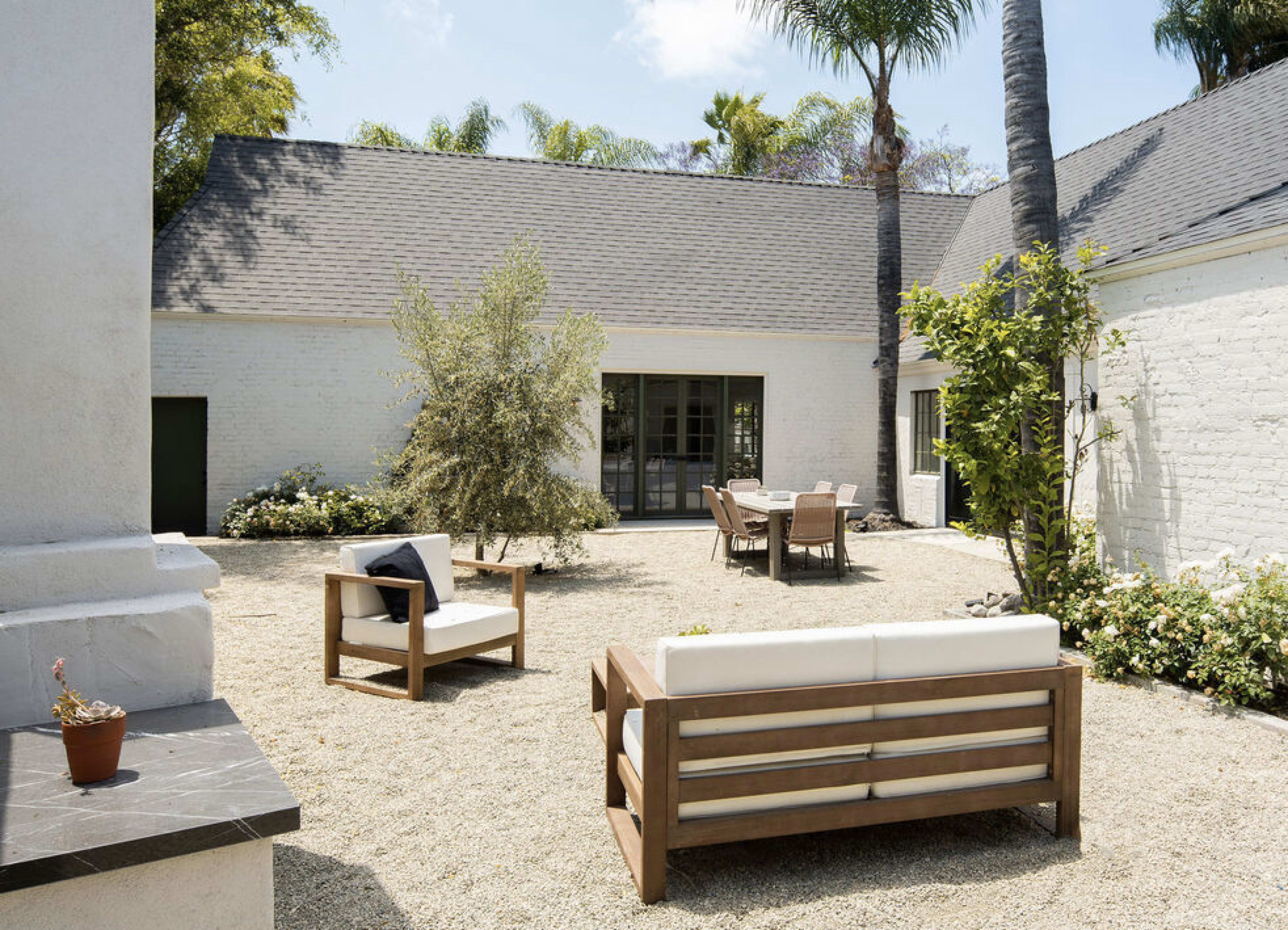 A minimalist outdoor patio features wooden furniture and a gravel surface, surrounded by greenery and a white brick building.