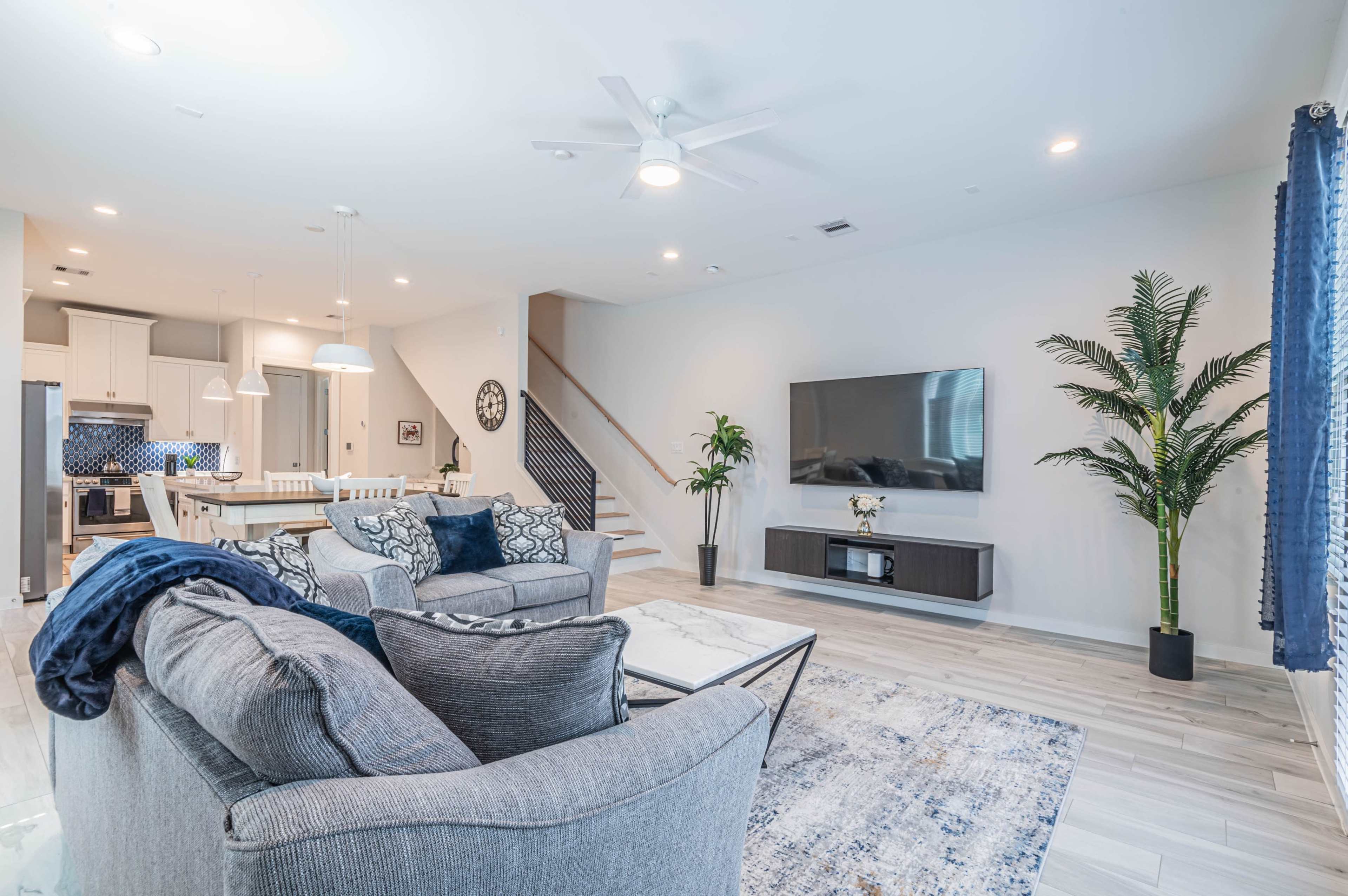 A modern living room featuring two gray sofas, a large flat-screen TV mounted on the wall, and a dining area in the background with white furniture and a blue accent wall.