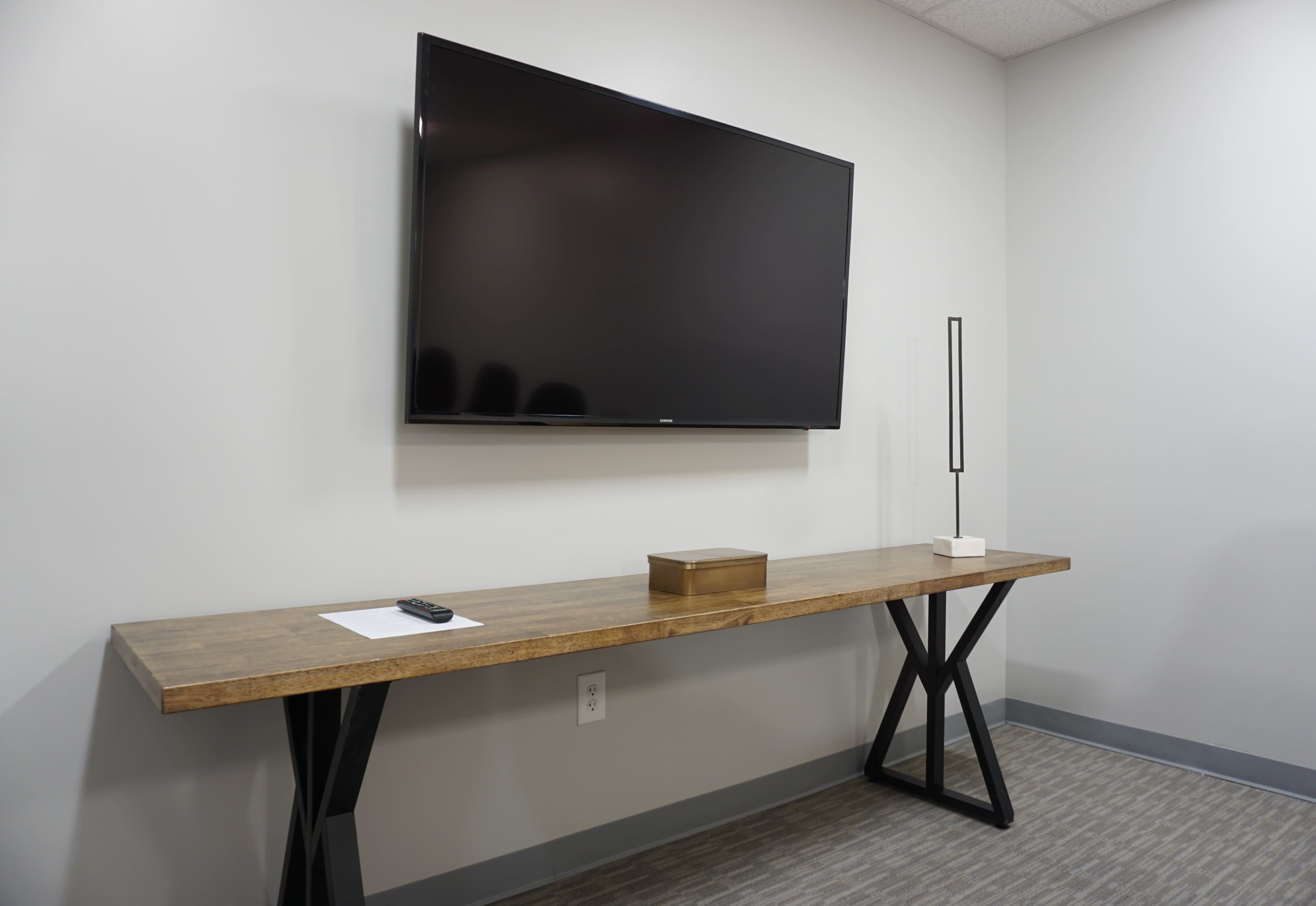 The image shows a minimalist room with a mounted flat-screen TV above a long wooden table, accompanied by a lamp and a decorative box.