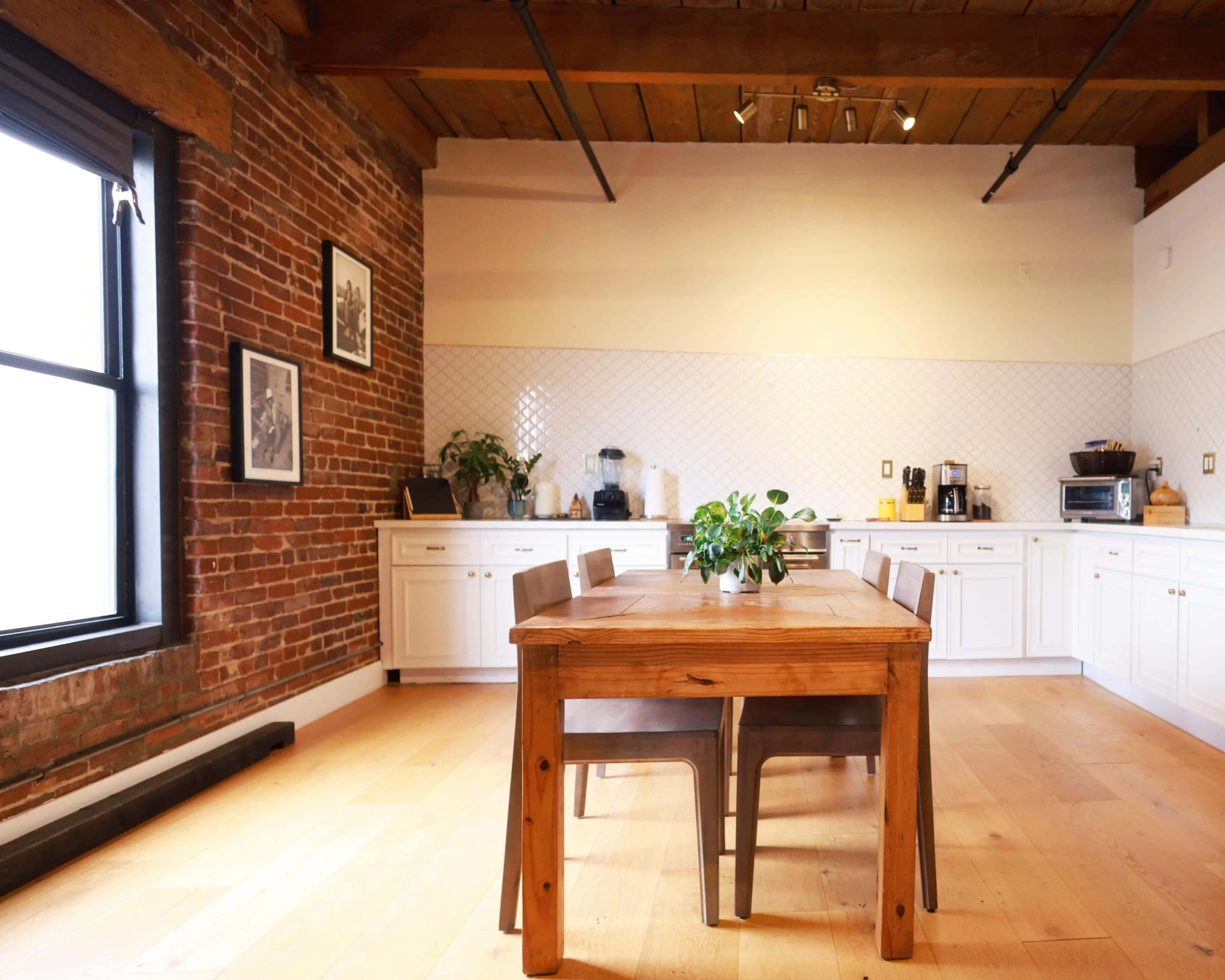A rustic kitchen features a wooden dining table surrounded by chairs, with exposed brick walls and white cabinetry.