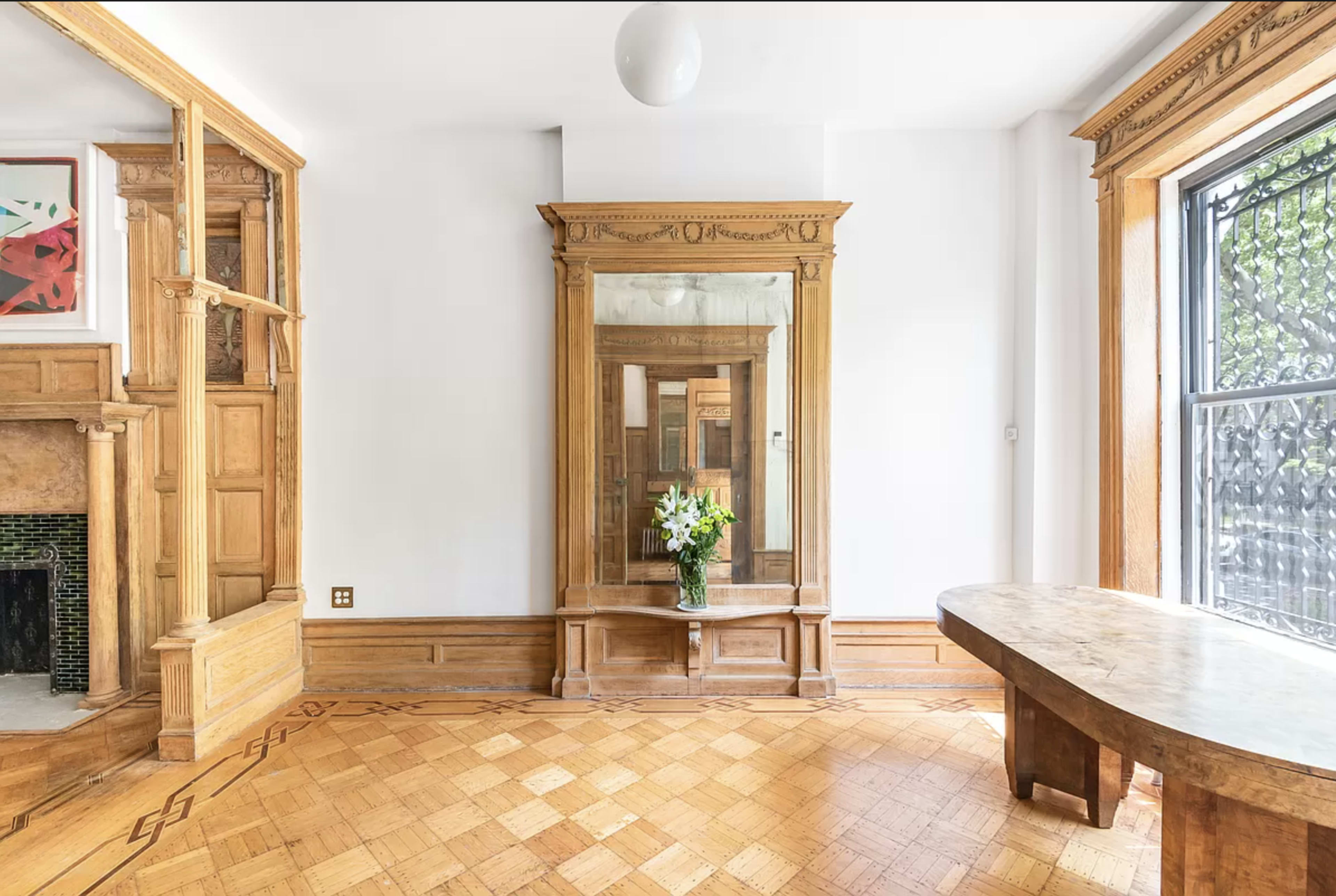 The image shows an empty room featuring a large wooden mirror, a floral arrangement on a wooden table, and intricate parquet flooring.