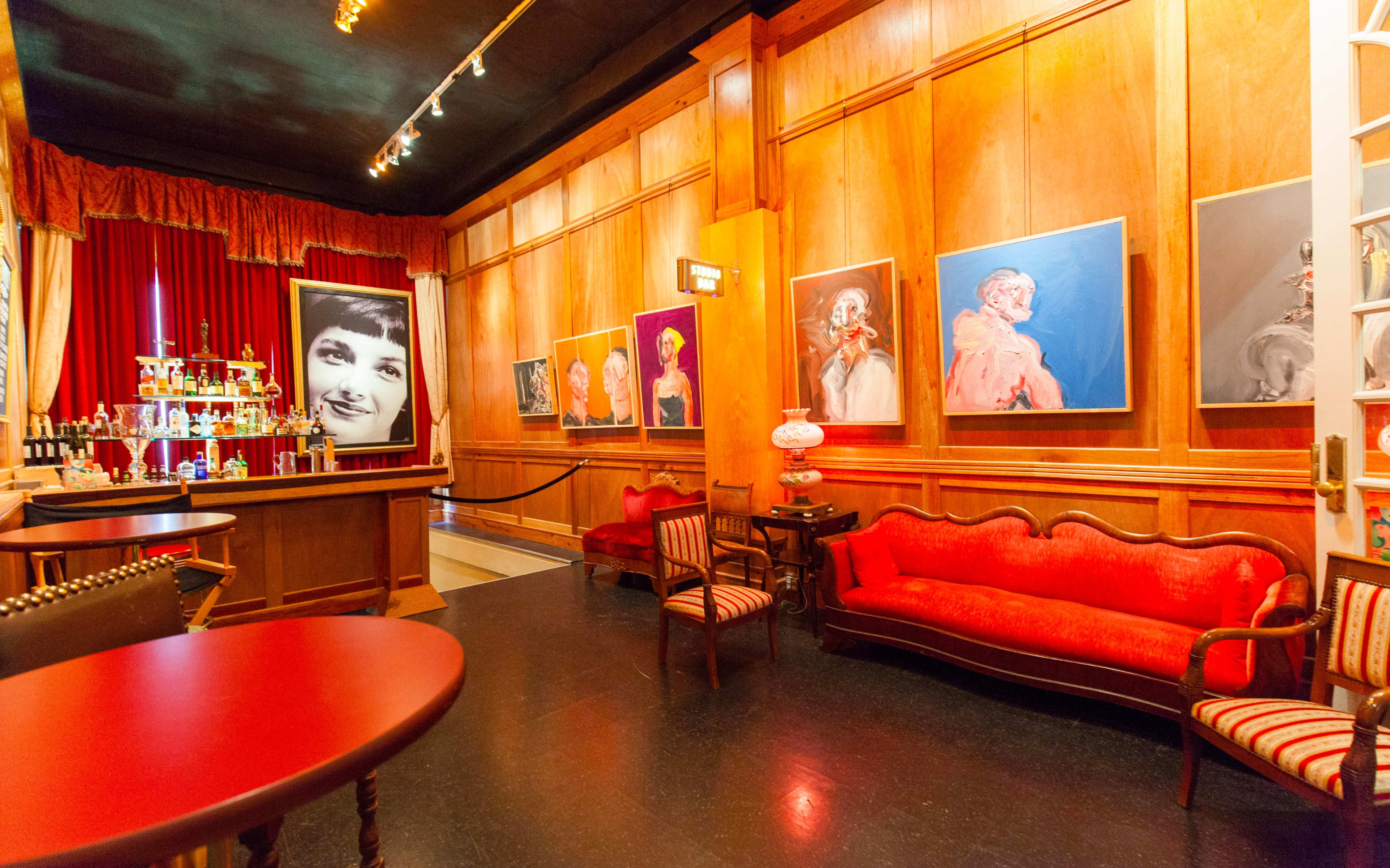 A warmly lit interior featuring a bar with a selection of bottles, red curtains, wooden walls, and large framed portraits on display.