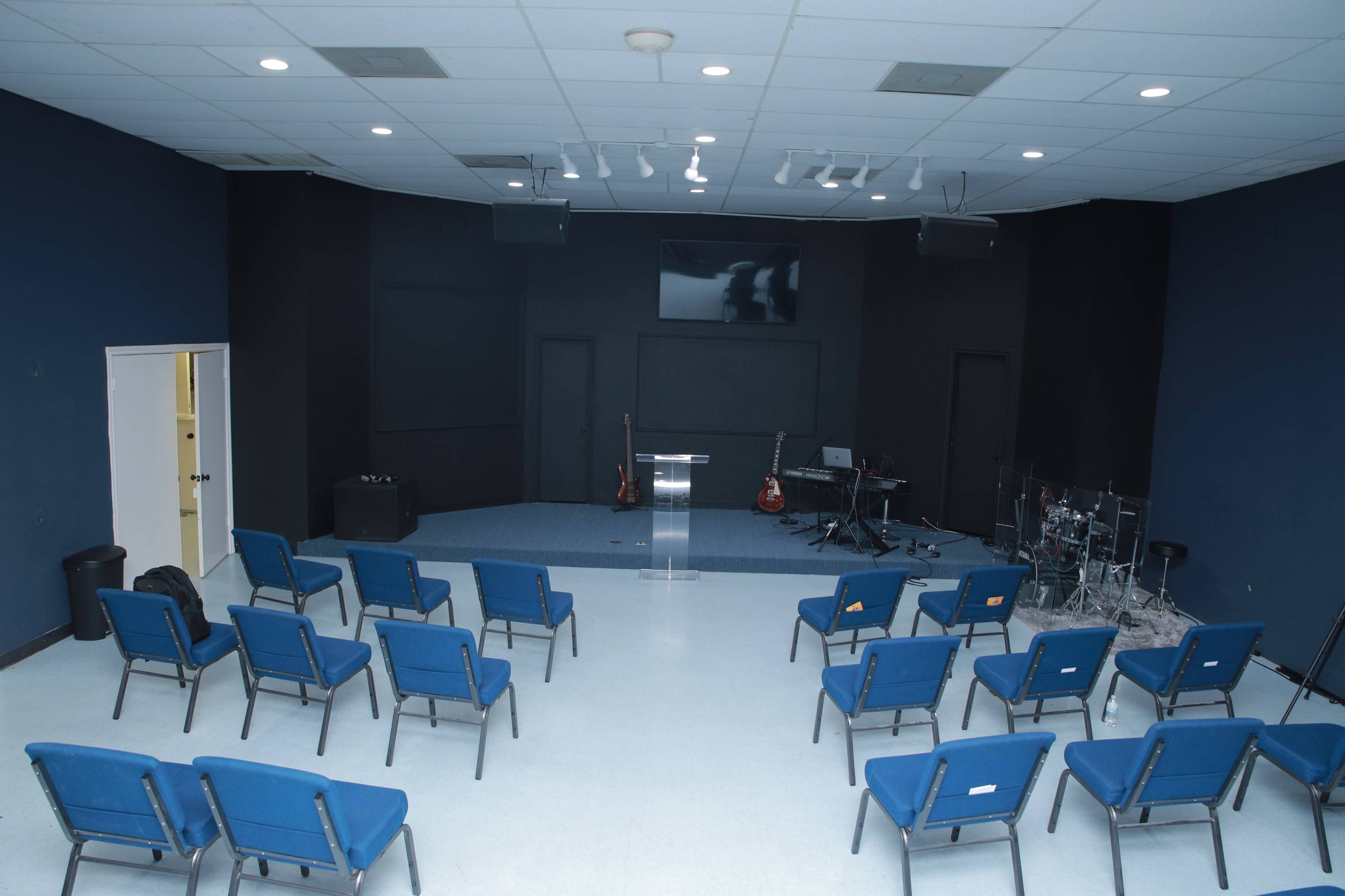 A room set up for a gathering, featuring rows of blue chairs facing a stage with instruments and a projection screen.