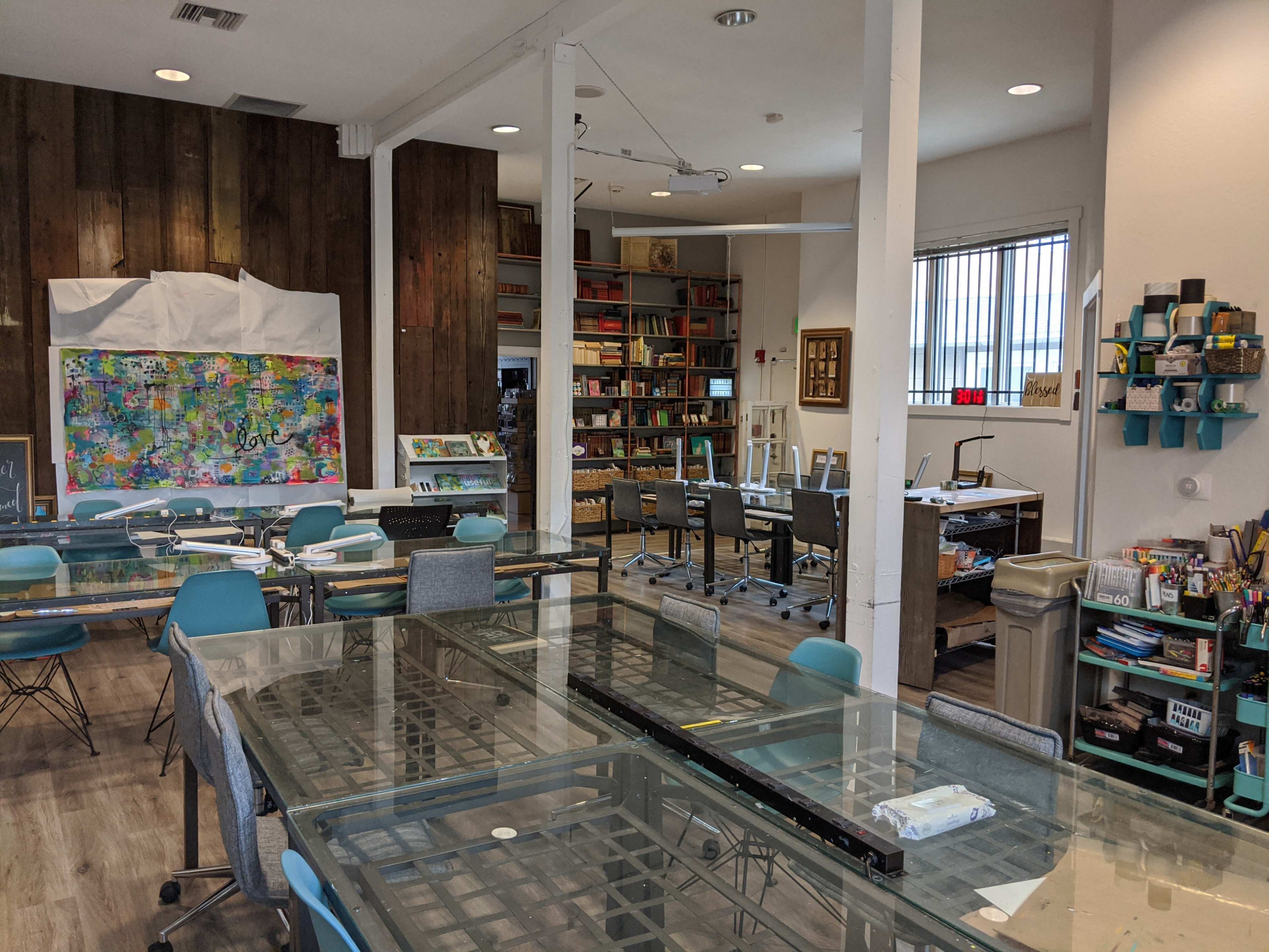 The image shows a bright art studio with tables, chairs, and shelves filled with various art supplies and books.