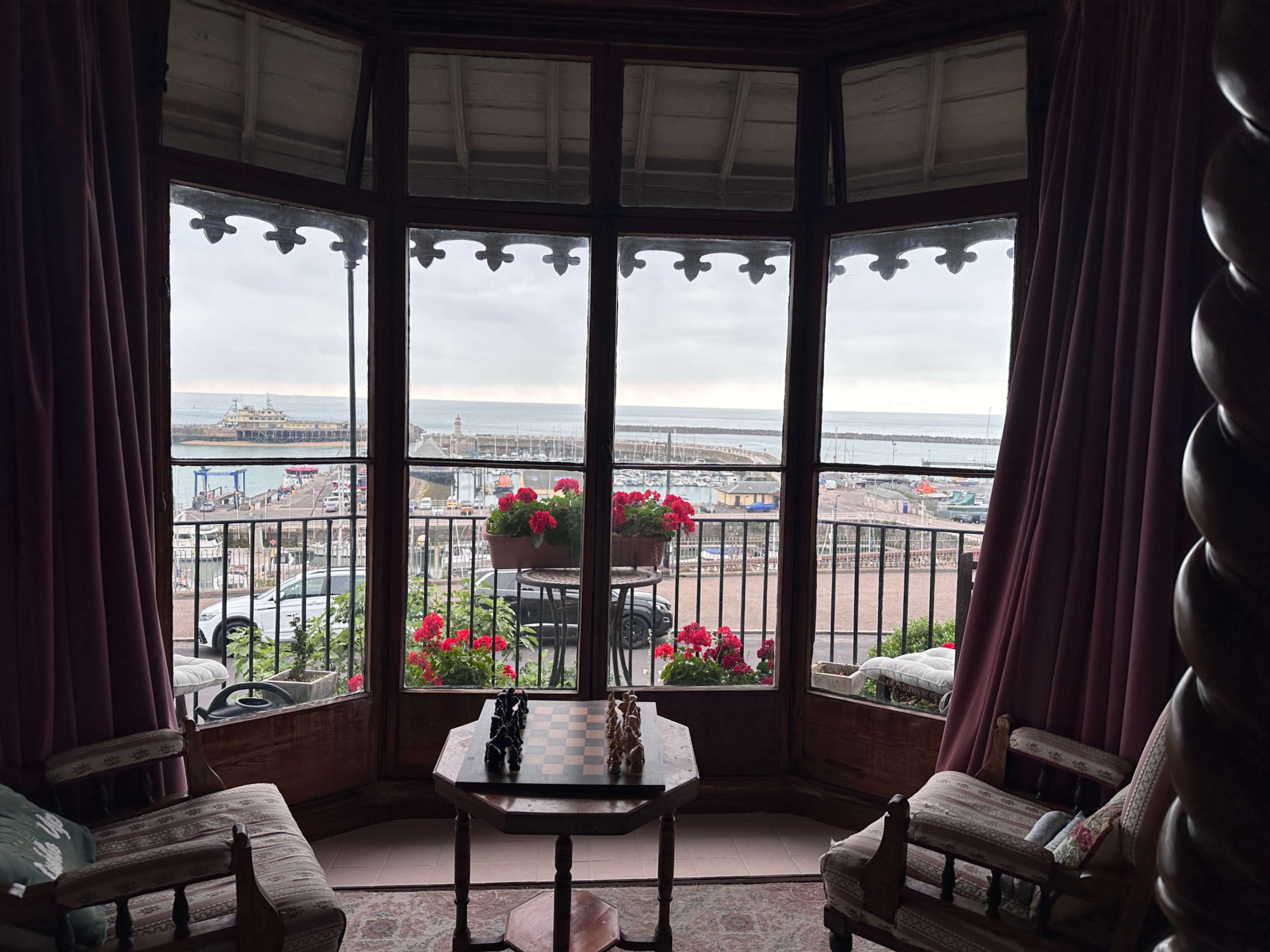 The image shows a cozy room with large windows overlooking a harbor, featuring a chessboard and two chairs.