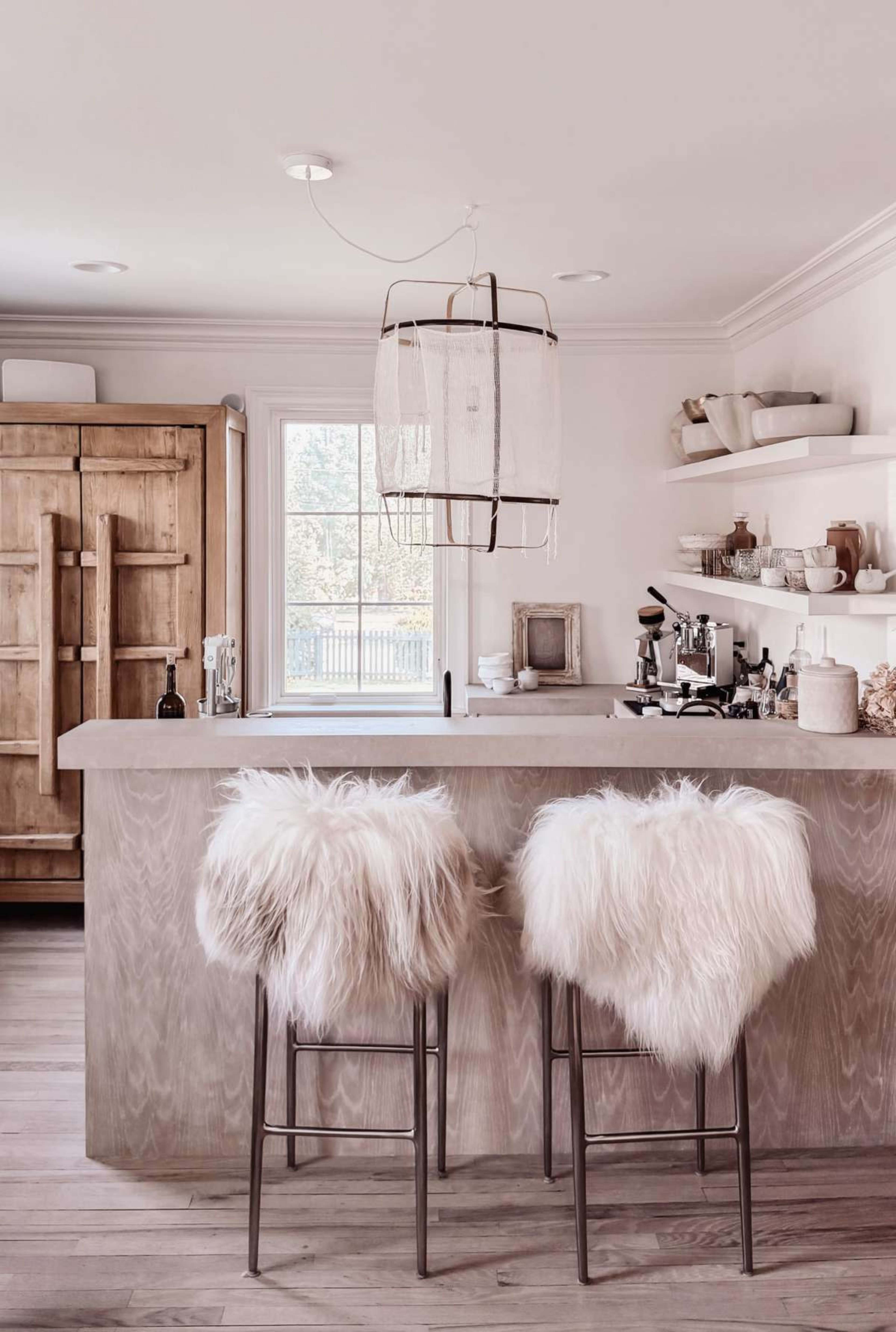 The image shows a modern kitchen with a light wooden bar, two bar stools with fluffy white seat covers, and a large window providing natural light.